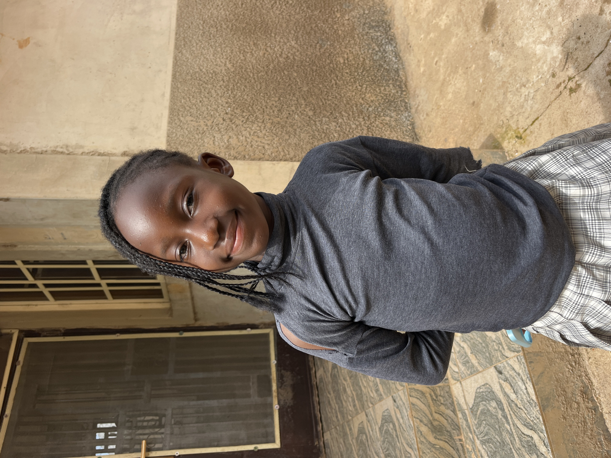A young Black girl is standing on a tiled staircase outside a building. She is looking towards the camera with a slight smile. She has braided hair pulled to the side and is wearing a dark gray turtleneck shirt with cutout shoulders and a black and white plaid skirt. Her hands are clasped behind her back. The building behind her is made of concrete and has a door with a screen and a window with panes. The ground is also made of concrete. The image appears to have been taken outdoors in a setting that is likely an urban or residential area.