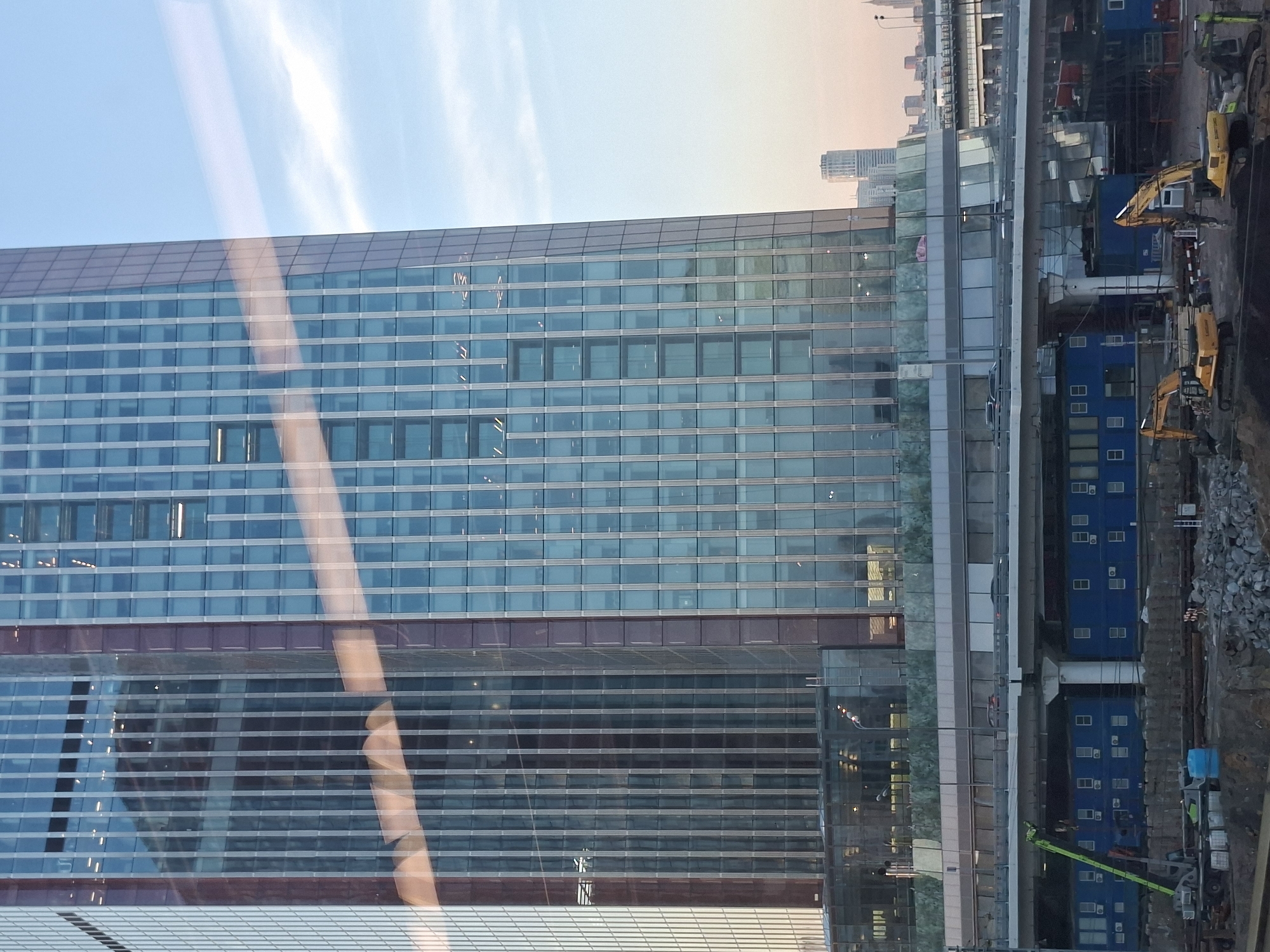 This is a medium shot of a modern skyscraper with a reflective glass facade. The building is tall and rectangular, with multiple floors visible. The glass reflects the sky and surrounding environment, creating a sense of depth and light. In the foreground, there is a construction site with heavy machinery, including excavators and a crane. There are also blue modular buildings, likely temporary structures for the construction crew. A road or bridge runs across the middle ground, with cars visible on it. The sky is a light blue with some clouds, indicating it is daytime. The overall scene suggests urban development and construction in progress.