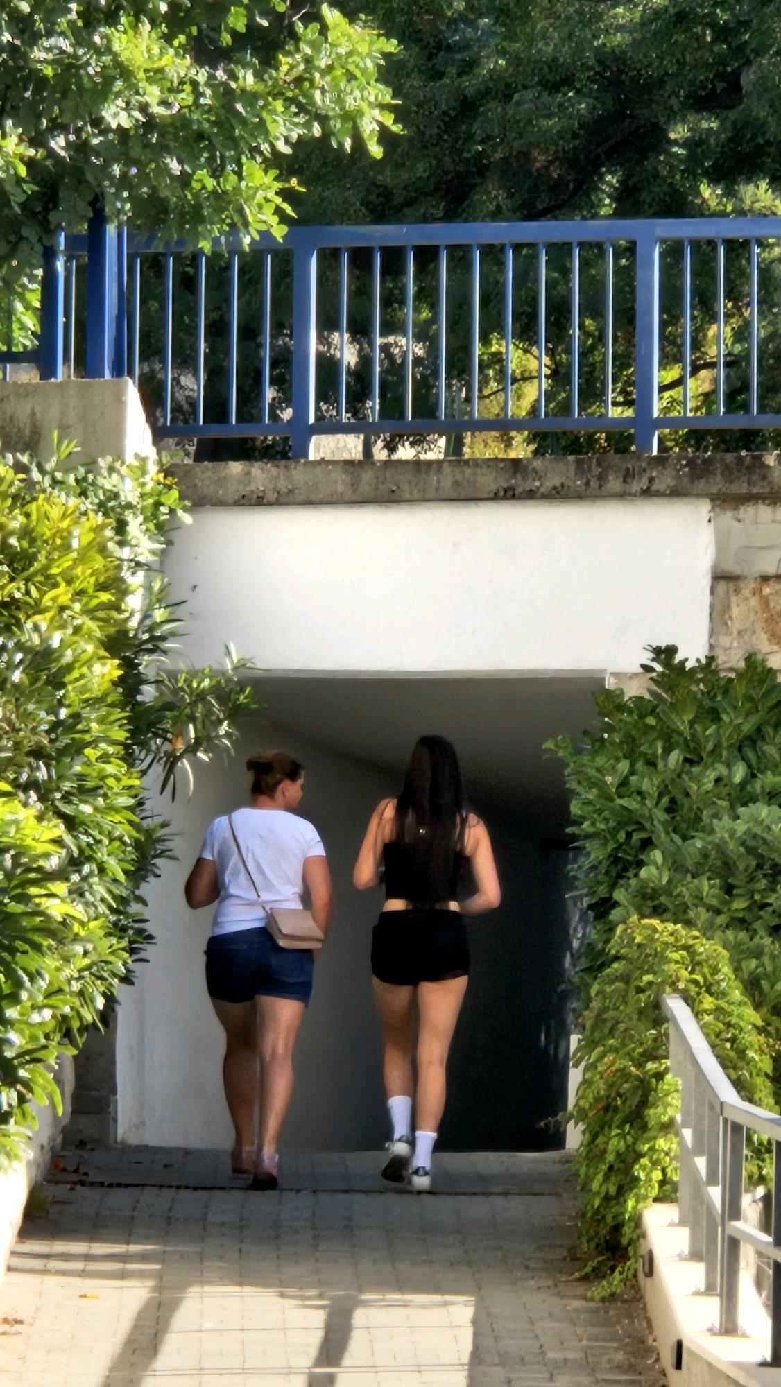 The image depicts two individuals walking down a paved pathway towards an arched entryway, framed by lush green foliage on both sides. The pathway is illuminated by dappled sunlight, casting shadows. A blue railing runs along the upper left portion of the scene, set against a backdrop of trees and a stone wall.
The person on the left is wearing a white t-shirt, denim shorts, and carrying a small light brown crossbody bag. Their hair is pulled back in a bun. The person on the right has long dark hair, is wearing a black crop top and black shorts, with white socks and athletic shoes. Both individuals are walking away from the viewer and towards the entryway. The entryway appears to be a tunnel or covered passage, with a stark white wall visible inside.
The location is identified as Općina Tučepi, Croatia. The architecture and vegetation suggest a Mediterranean setting. The overall scene conveys a sense of movement and outdoor activity.