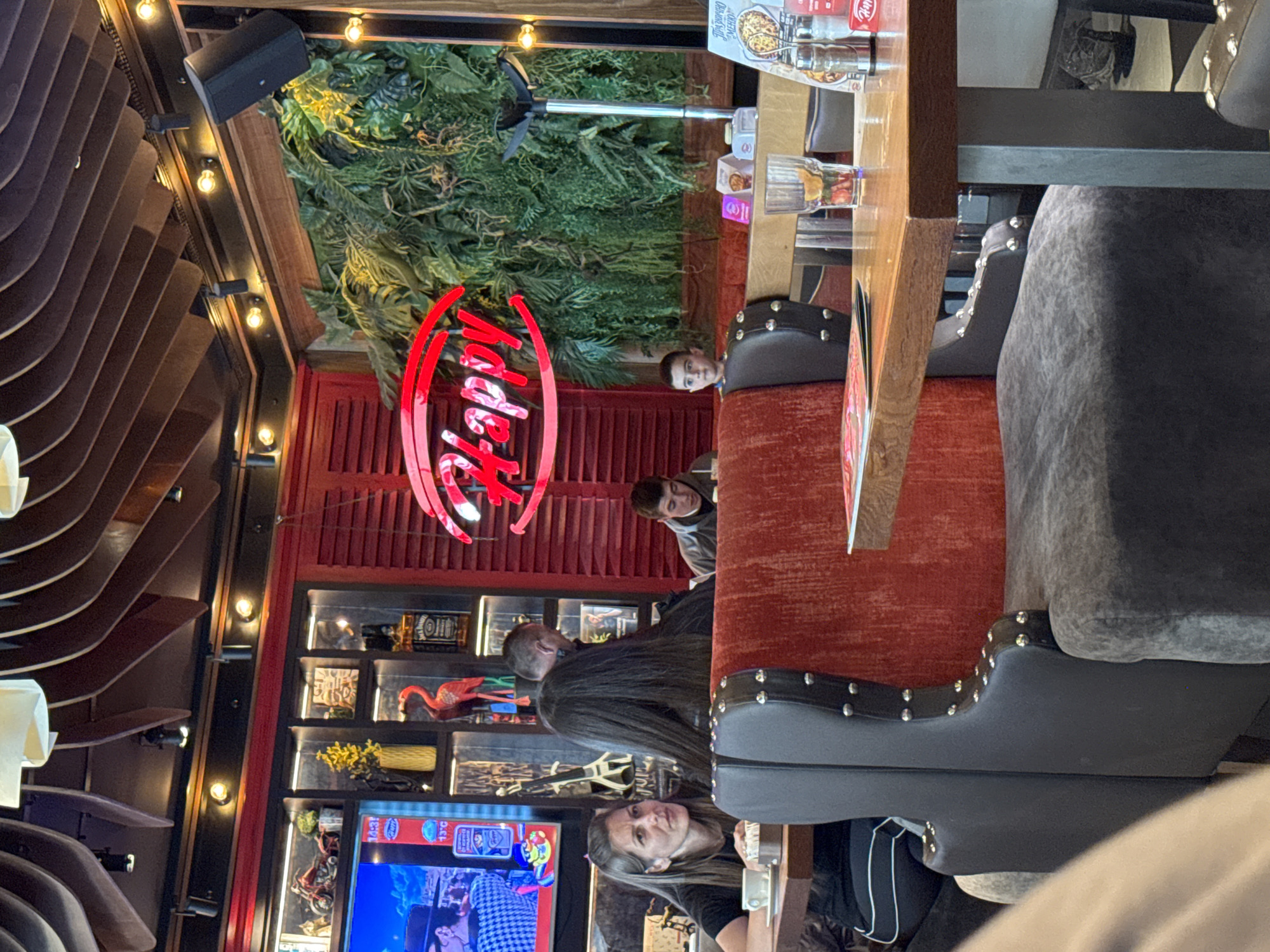 The image shows the interior of a restaurant, likely in Plovdiv, Bulgaria, given the signage in Cyrillic. People are seated at tables, with a woman visible in the foreground. A TV is visible in the background and shelves are decorated with various objects. Overhead, there are decorative plants and a speaker. The restaurant is decorated with red colors.