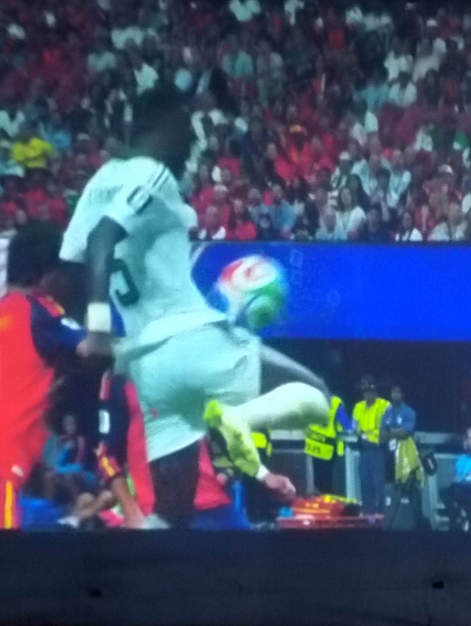 This is a blurry, pixelated image of a soccer match. A player in a white jersey with black stripes is in mid-air, appearing to kick a ball. Another player in a red and blue uniform is to his left. Spectators are visible in the background, many wearing red. The scene appears to be on a grass field, likely a stadium. The image does not provide any information about the city of Payagaboh, Indonesia.