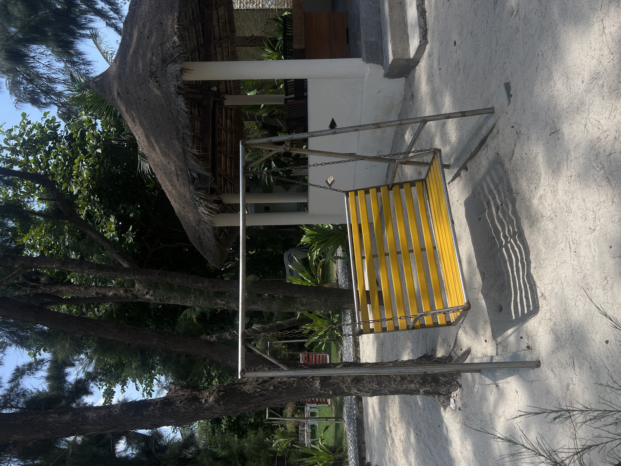 A yellow swing set rests on white sand next to a building with a thatched roof, located in Dong Ninh Hoa Ward, Vietnam. Trees surround the area.