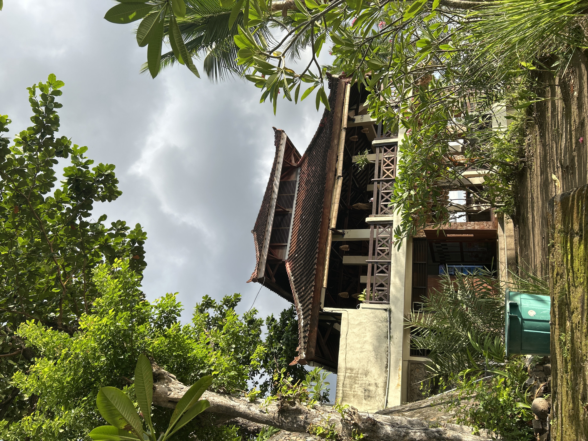 The image depicts a building with a multi-tiered roof in Senggigi, Indonesia. The structure is surrounded by lush greenery, including various trees and plants. The sky is overcast with clouds. The building appears to have a beige exterior with wooden balcony railings. A green container is visible near the base of the building on the right. The perspective is from a low angle, looking upwards.