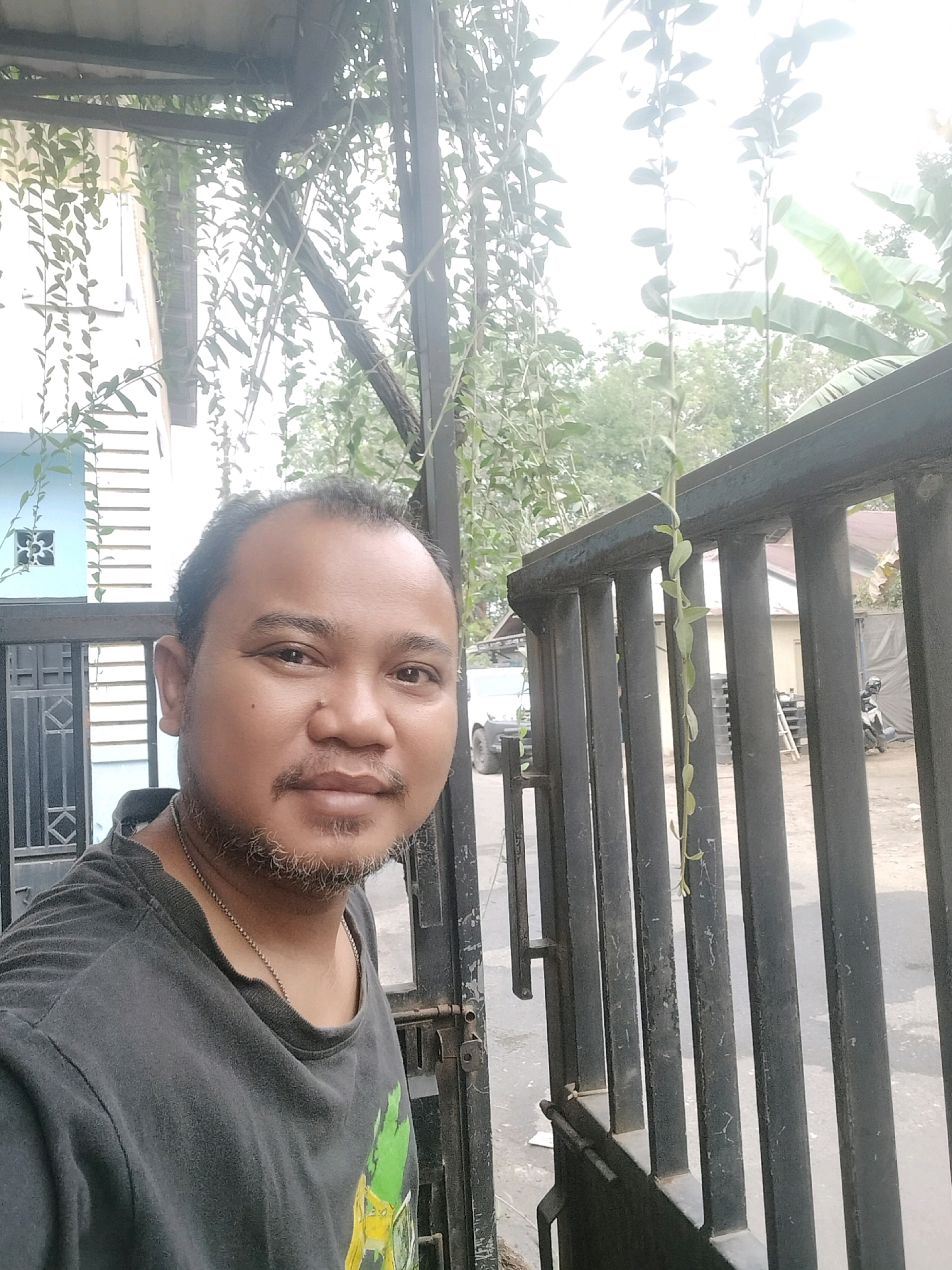 A medium shot captured from a low angle shows a man with dark skin, a receding hairline, and a goatee, standing outdoors in front of a black metal fence. He is wearing a dark gray t-shirt with a green and yellow graphic on it and a silver chain. The man is looking directly at the camera with a neutral expression. Behind him, a light blue house with white trim is visible. Green vines hang from a corrugated metal roof structure overhead, and trees and banana leaves are in the background. A motorcycle is parked on the right side of the image. The scene appears to be in Pembataan, Indonesia, given the presence of traditional housing and tropical vegetation. The lighting suggests it is daytime, with the sky being overcast or very bright.