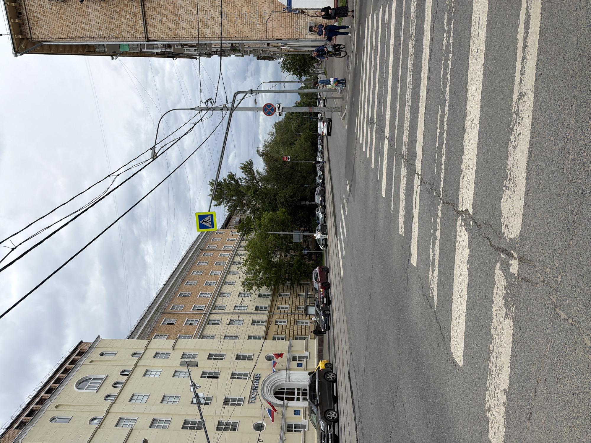 A street scene in an urban environment under an overcast sky shows a pedestrian crossing in the foreground. The crossing is marked with white stripes on asphalt, with cracks visible in the road. Across the street, several cars are parked or driving. On the left side of the street is a large, multi-story building with a light yellow facade, featuring numerous windows and an arched entranceway with flags. On the right side of the street, a brick building of similar height is visible. People are seen on the sidewalk and near the crossing. Traffic lights and overhead wires are present, indicating the presence of infrastructure. Trees line the side of the road, and more buildings are visible in the background. The overall atmosphere is that of a typical city street on a cloudy day.