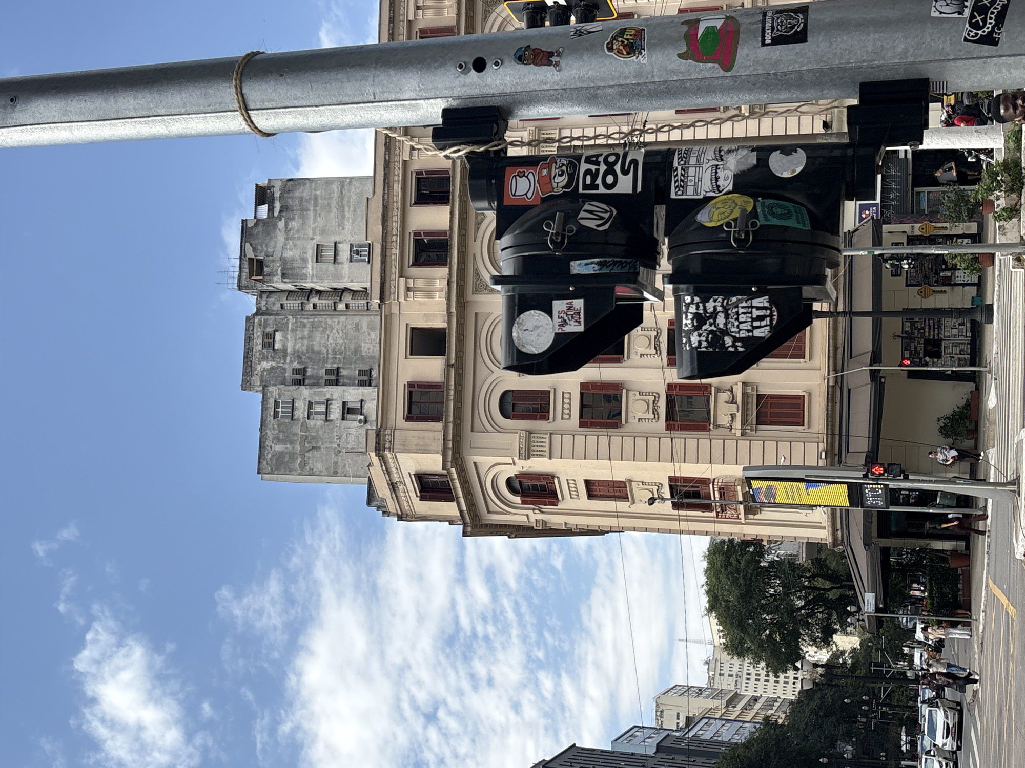 A street scene in São Paulo, Brazil, features a traffic light pole in the foreground adorned with stickers. Behind it, a pale yellow building with red-shuttered windows rises, topped by a gray, Brutalist-style building. The sky above is blue with scattered white clouds. On the street level, pedestrians walk, cars are visible, and a digital display shows "31°C". The image captures a typical urban environment with a mix of architectural styles and everyday city life.