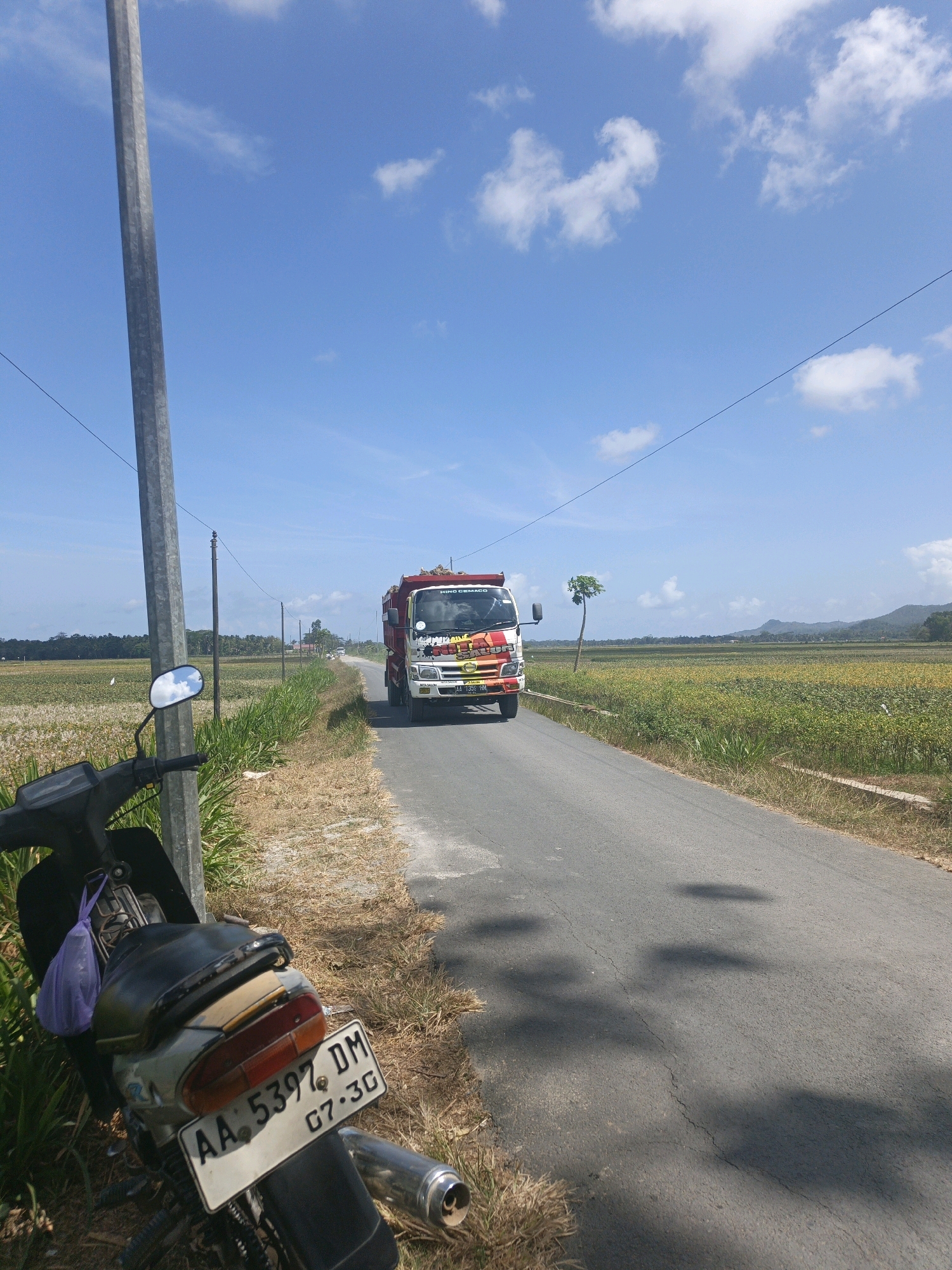 The image depicts a rural road scene in Kamulyan, Indonesia, under a bright blue sky dotted with scattered white clouds. In the foreground, to the left, a motorcycle is parked next to a tall, grey utility pole. The motorcycle is dark-colored with a visible license plate displaying "AA 5397 DM 07-30". A purple plastic bag is attached to the back of the motorcycle. The road itself is paved and appears to be in moderate condition, with some cracks visible. To the right of the road, fields of green and yellow vegetation stretch into the distance, bordered by a treeline. Further down the road, a red and white truck is traveling away from the viewer. The truck appears to be loaded with goods. The overall environment suggests a sunny day in an agricultural area.