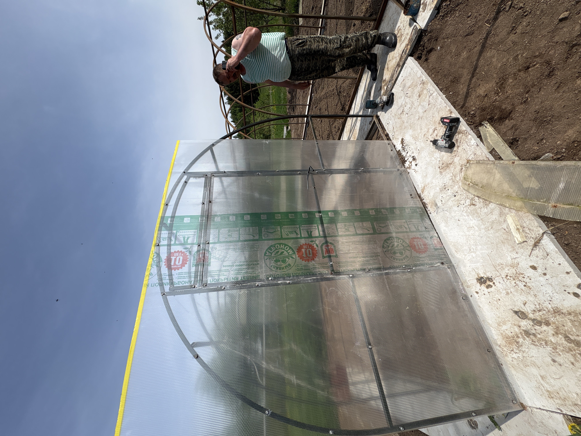 A man is building a greenhouse in Lukintsy, Russia. The structure is made of metal and plastic panels. The man is wearing a striped shirt and camouflage pants. Tools are visible near the construction site. The background consists of a partially clear sky.