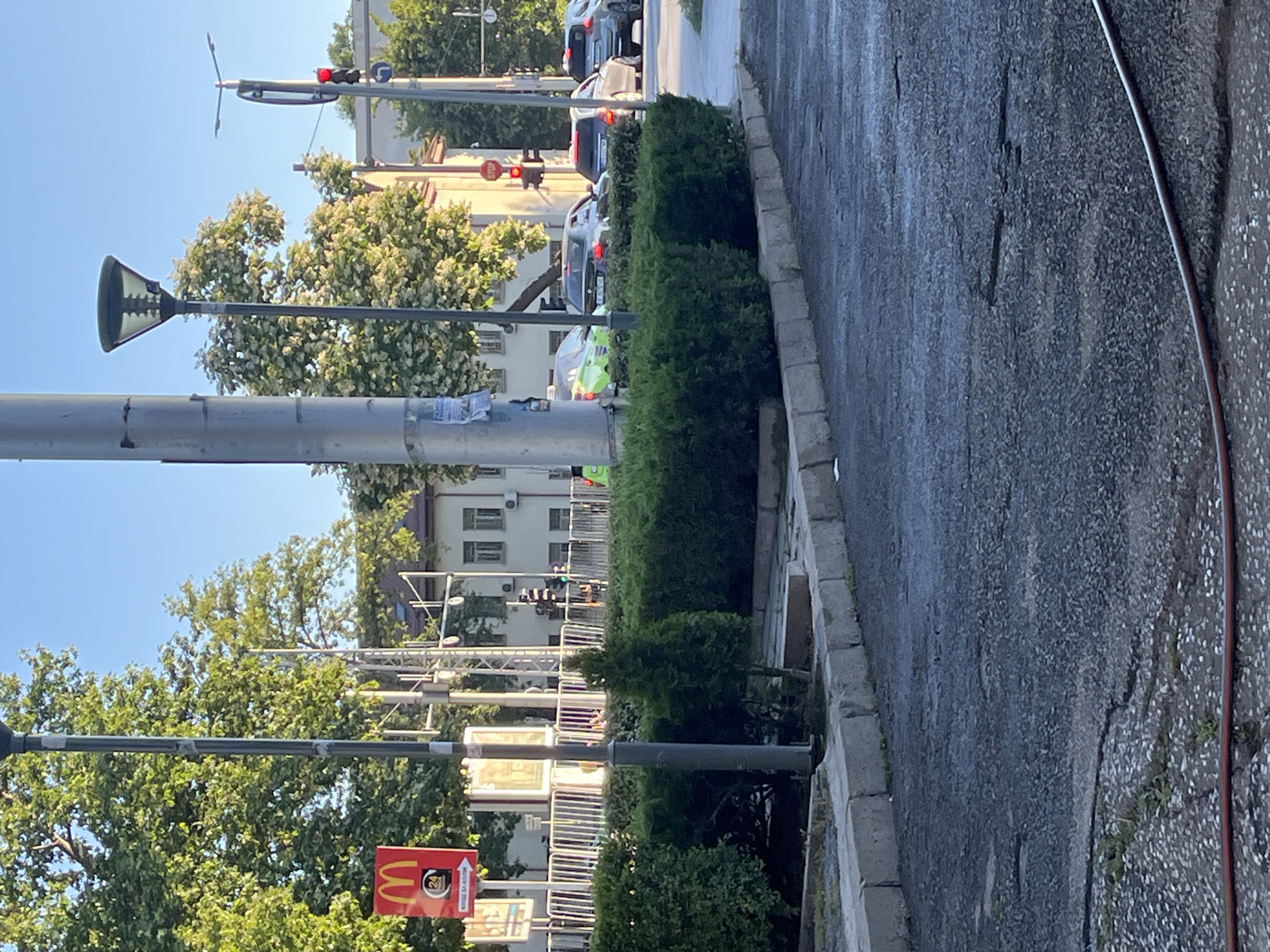 The image depicts a street scene in Plovdiv, Bulgaria, likely near a McDonald's restaurant, as indicated by a visible red sign with the McDonald's logo and "24 HOURS" text. The photograph captures a street with cars, a traffic light showing red, and lush green trees and hedges. A lamppost stands prominently in the foreground, and a metallic fence borders a pedestrian walkway. The road surface appears wet, suggesting recent rain or cleaning. In the background, buildings with white facades and multiple windows are visible. The sky is clear and blue, indicating a sunny day.