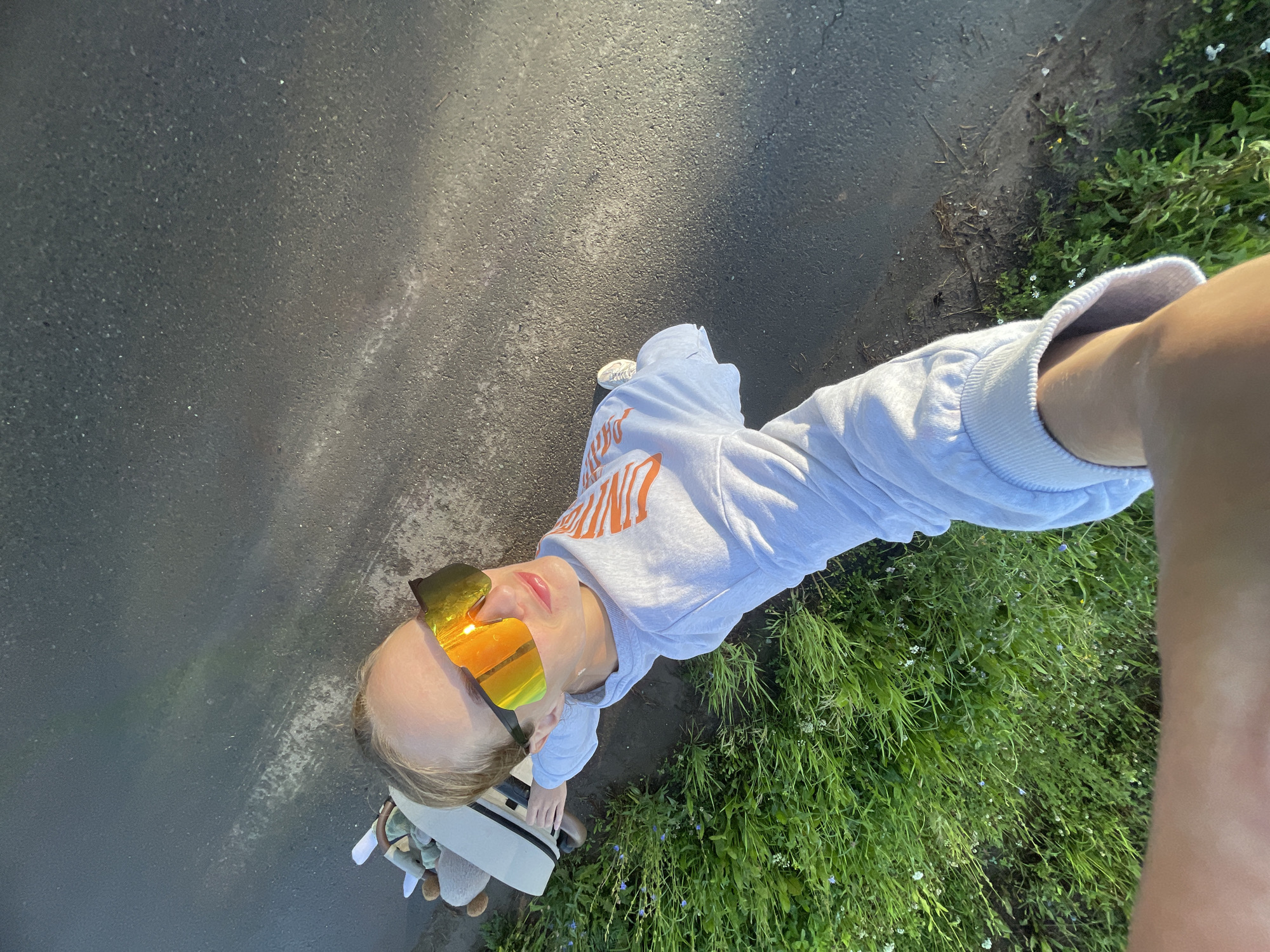 A person is standing on asphalt, near a patch of grass with small white and blue flowers. The person is wearing a light gray hoodie with orange lettering on the sleeve, and white sneakers. They have large, reflective orange and yellow sunglasses on. A light-colored stroller, possibly with stuffed animals visible, is to the left of the person. The ground is dark asphalt with some white markings. The scene is outdoors, with natural lighting. The location is identified as Podshivalovo, Russia.