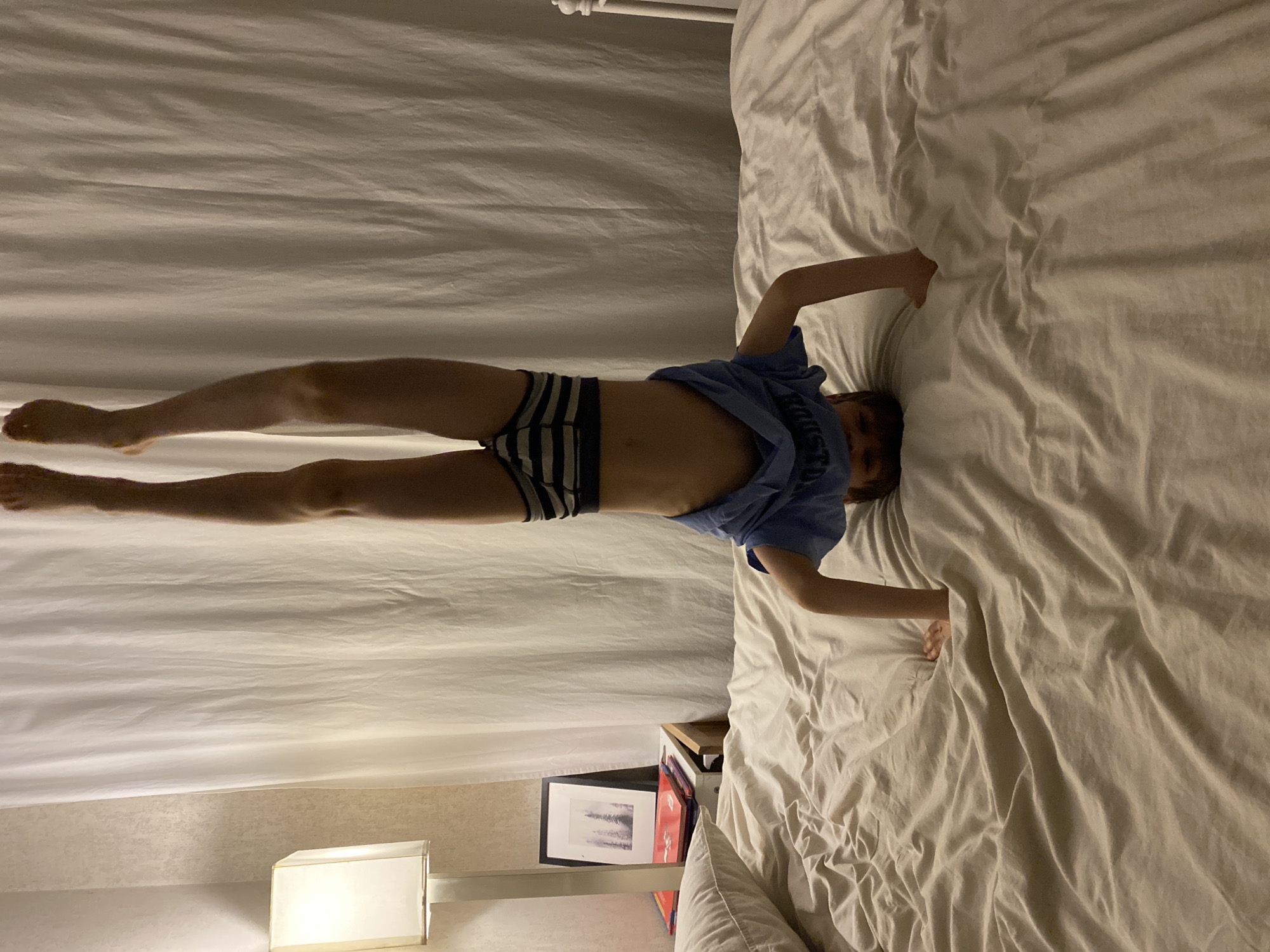 A child lies on a white bed in a room with a lamp and a nightstand. The child is wearing striped underwear and a blue t-shirt. There is a picture on the nightstand. No information suggests the location is Yekaterinburg, Russia.