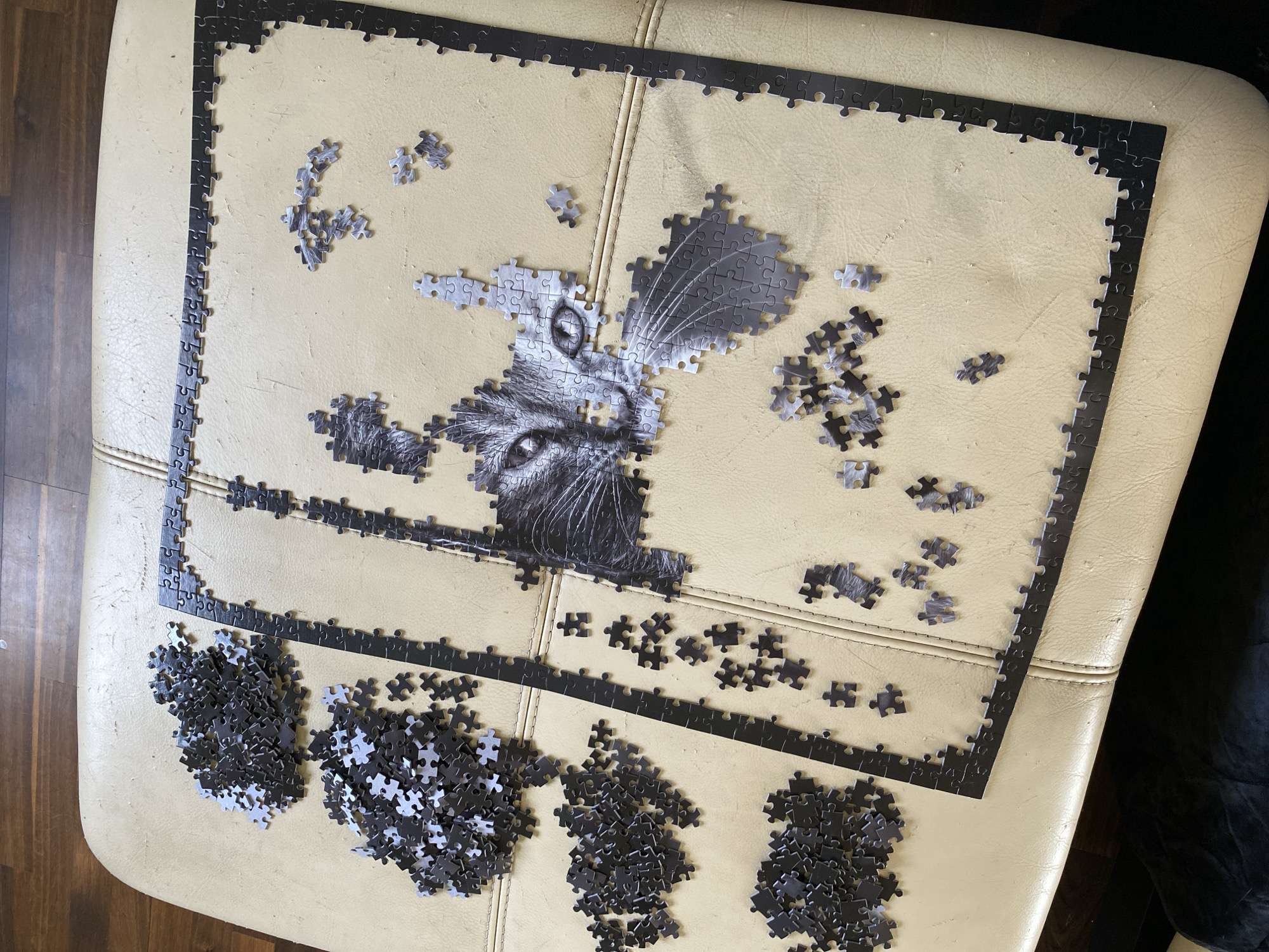 A jigsaw puzzle of a cat is partially assembled on a beige ottoman. The puzzle's border is complete. Some of the cat's head is assembled in the center of the puzzle, with piles of unassembled pieces surrounding it. The ottoman is located on a wooden floor, near what appears to be a dark-colored piece of furniture. The location is not discernable as Villaviciosa, Spain, from the given image.