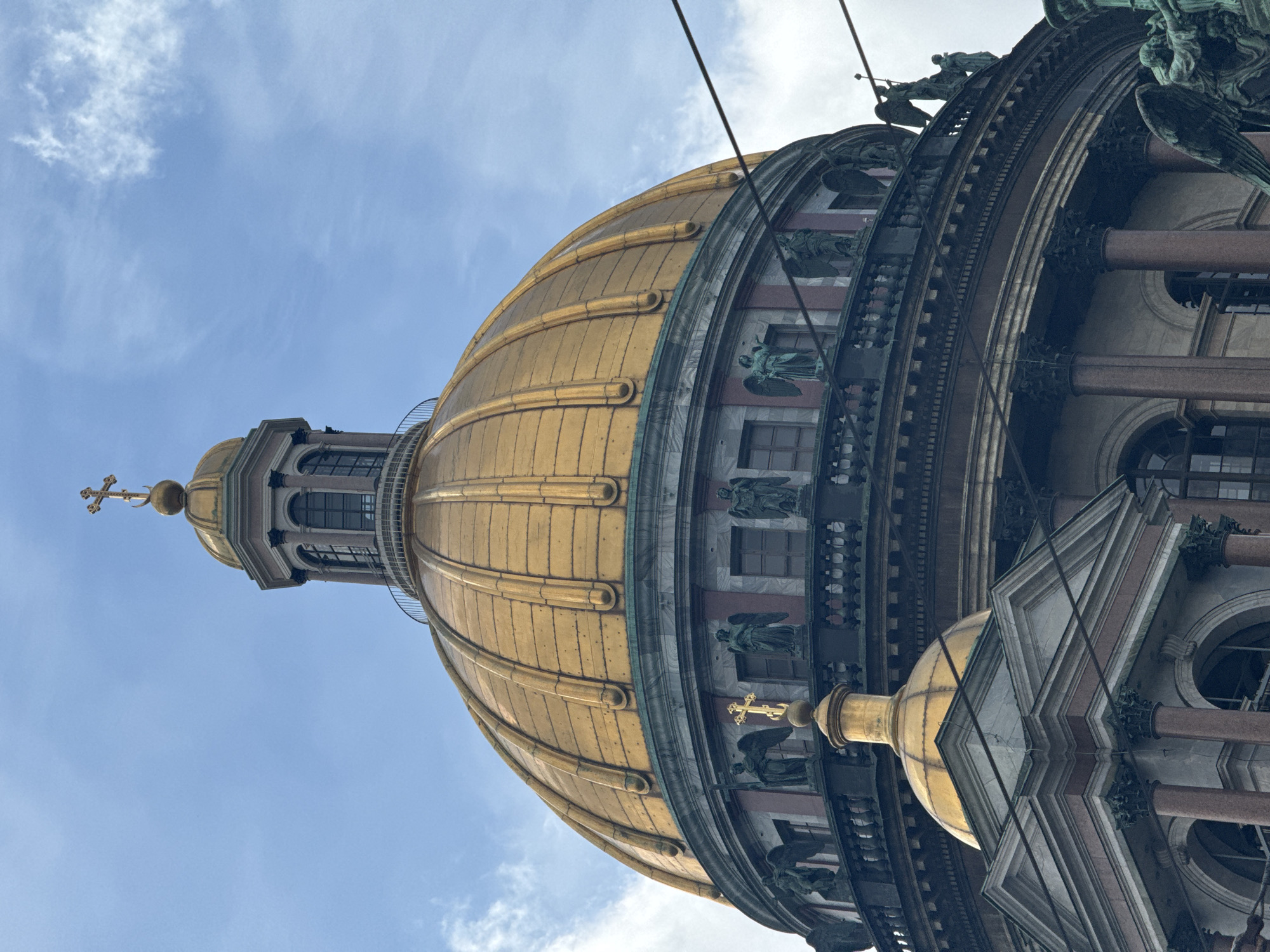 A low-angle, close-up shot captures the majestic golden dome of Saint Isaac's Cathedral in Saint Petersburg, Russia, against a bright blue sky with scattered white clouds. The gilded dome dominates the frame, featuring distinct ribs that run from its base to the apex. At the very top, a smaller cupola supports a prominent golden cross.
Below the main dome, the drum of the cathedral is visible, adorned with intricate architectural details, including several windows, ornate friezes, and numerous sculptures. These statues depict figures, including winged angels, and appear to be made of bronze, showing signs of patina with their greenish hue. Robust, reddish-brown granite columns are also visible, supporting sections of the structure. Thin, dark wires or cables stretch across the upper portion of the image, partially obscuring the dome and sky. The scene is brightly lit, indicating a clear, sunny daytime. There is no visible text in the image.