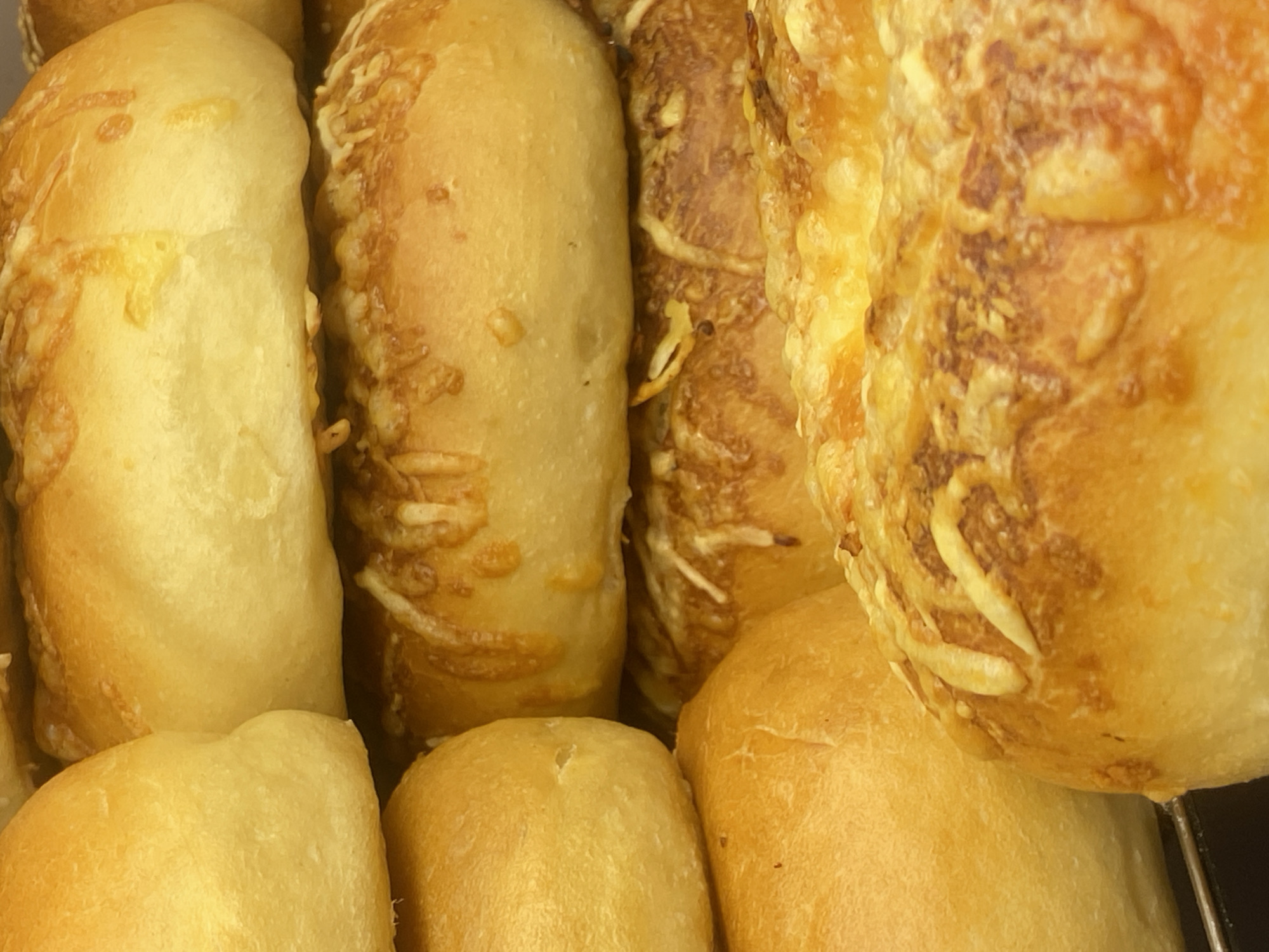 A close-up, high-angle shot shows a stack of golden-brown baked goods, possibly bread rolls or buns, some topped with melted cheese. The texture of the baked goods suggests they are fresh. The background is blurred, obscuring any details about the environment. There are no people or specific landmarks visible in the image that would allow for the determination of a location in Warsaw, Poland, or any other city.