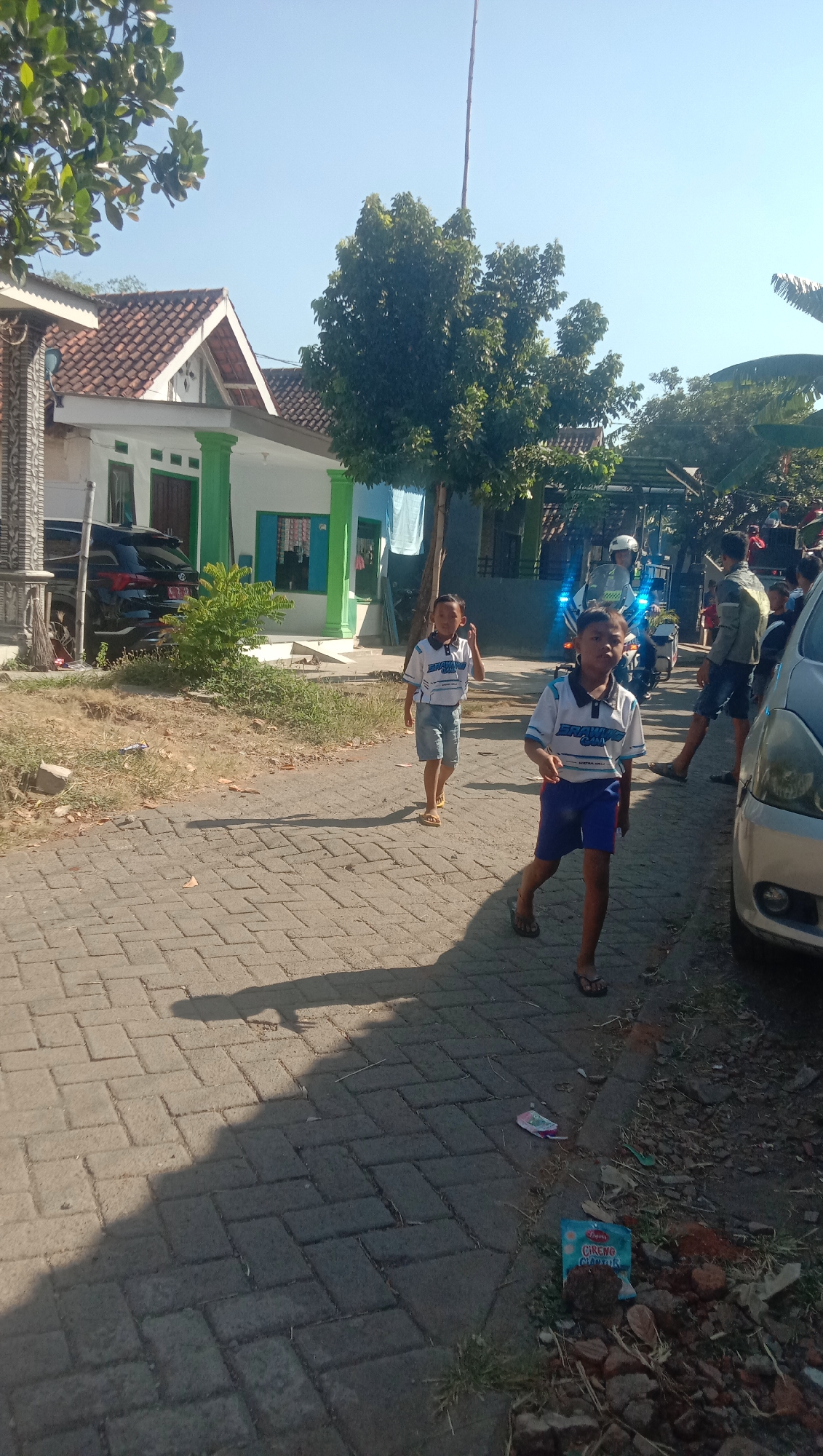 The image depicts a street scene in Ngempit, Indonesia, under a clear blue sky. Two young boys, one in the foreground and another further back, are walking on a paved street. Both are wearing matching blue and white striped shirts and denim shorts. The boy in the foreground is looking directly at the camera, while the boy behind him is looking to his left.

Behind the boys, a police officer on a motorcycle with flashing blue lights is visible, suggesting some activity or event is taking place. Several other people are present in the background, some standing and others walking, but their details are indistinct. A silver car is parked on the right side of the street.

The environment shows residential buildings with tiled roofs and green painted columns. Trees and foliage are scattered throughout the scene. The street is paved with interlocking brick pavers, and there is some dry grass and debris along the side of the road, including a discarded packet of "CIRENG CRISPY". Shadows from trees and people are cast on the street.