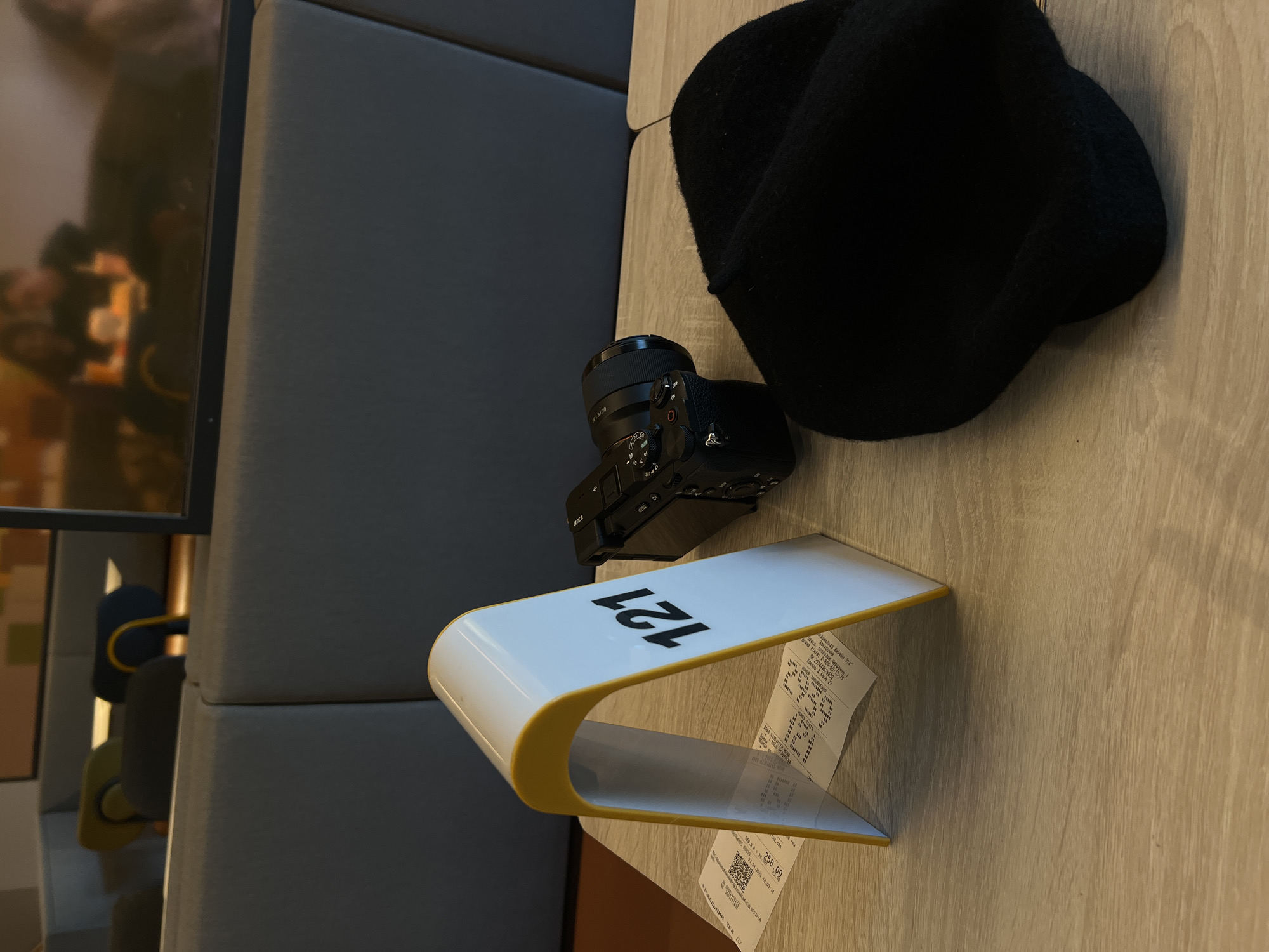 An indoor, slightly overhead shot captures a light-colored wooden table in what appears to be a cafe or dining area. On the table, a black digital camera, identifiable as a Sony Alpha series with an FE 1.8/50 lens, is positioned next to a black beanie or hat. A distinctive white, U-shaped stand with yellow trim displays the number "121" in black lettering. A crumpled paper receipt or check is tucked underneath this stand.
The background on the left shows a segment of a grey upholstered booth or bench. Further left, there are blurred reflections on a shiny surface, possibly a window or a dark, reflective wall, showing indistinct shapes that might be other patrons or parts of the establishment.
The visible text on the receipt confirms a Ukrainian setting. It includes a hotline number "0-800-50-15-79," a fiscal ID "Η 237444526652," and specifies "Ковель 1 Каса 29" (Kovel 1 Cashier 29). An amount of "2358.00" is clearly visible, followed by "ФІСКАЛЬНИЙ ЧЕK D" (Fiscal Check D). Other partially legible text includes "OS/813," "απι," and "Закусочив." The image depicts a casual scene of personal items on a table, likely shortly after a meal or purchase.