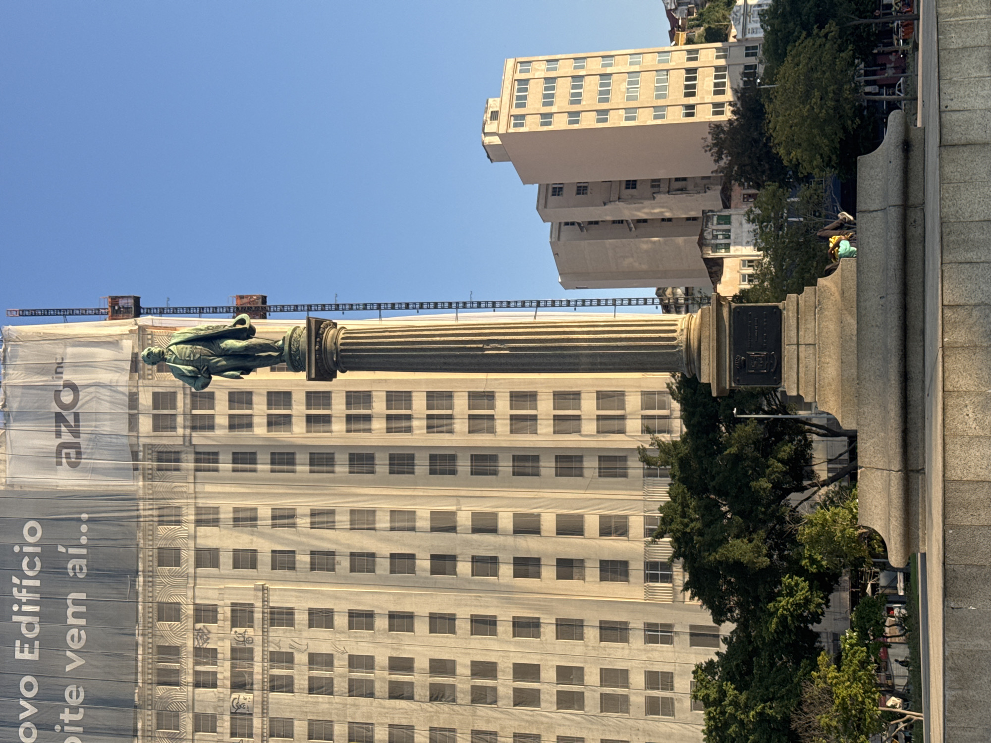 The image shows a statue on a column in front of a building undergoing renovation, with a large banner covering its facade. The banner has text in Portuguese: "Novo Edifício" and "site vem aí...". The sky is clear blue, suggesting a sunny day, likely in the morning or late afternoon given the shadows.
The statue depicts a person in a draped garment, possibly a historical figure, standing on a tall, fluted column. Behind the column and statue, the building is covered by a large white banner that obscures most of its architectural details. The banner is stretched taut and has some wrinkles, indicating it's a temporary covering. Through some sections of the banner, rows of windows are visible.
In the background, other buildings with many windows can be seen, suggesting an urban environment. Trees with green foliage are present, some taller than others, adding a natural element to the cityscape. There are also hints of human activity, with a few people visible in the distance, one possibly sitting on a stone structure. The overall scene appears to be a public space, likely a plaza or street in a city.
The perspective of the image is from a low angle, looking up at the statue and the buildings. The orientation of the image is rotated 90 degrees counter-clockwise. The scene is bright and appears well-lit, with the sunlight casting shadows. The presence of construction or renovation materials and the banner suggest that the building is either being built or refurbished. The clear sky and absence of rain indicate fair weather.