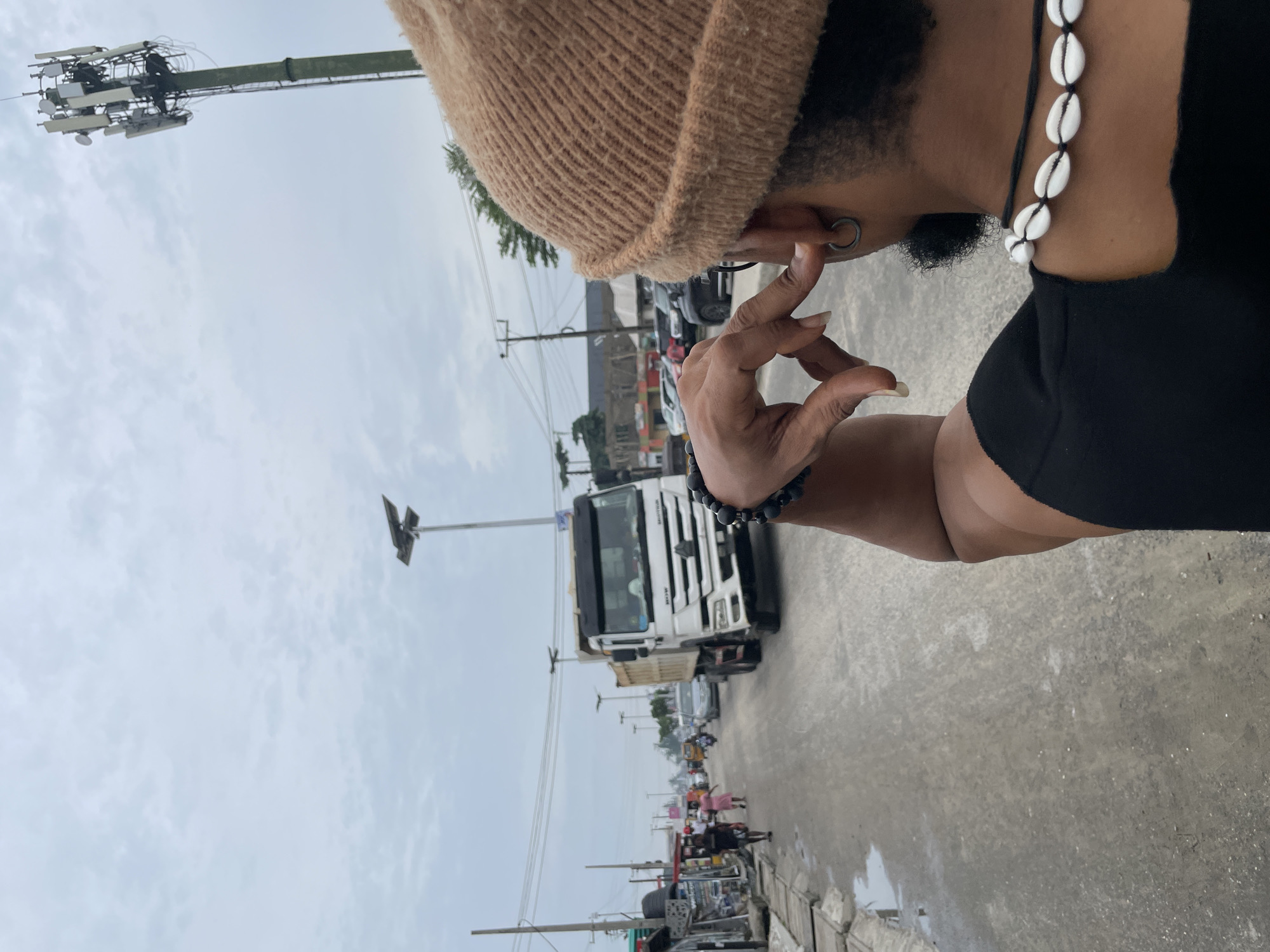 A street scene in Ado, Nigeria shows a person wearing a brown beanie, black top, cowrie shell necklace, and a beaded bracelet, standing near a road. A white truck is visible further down the road, along with other vehicles and pedestrians. Utility poles line the street, and a cell tower is in the background. The sky is overcast.