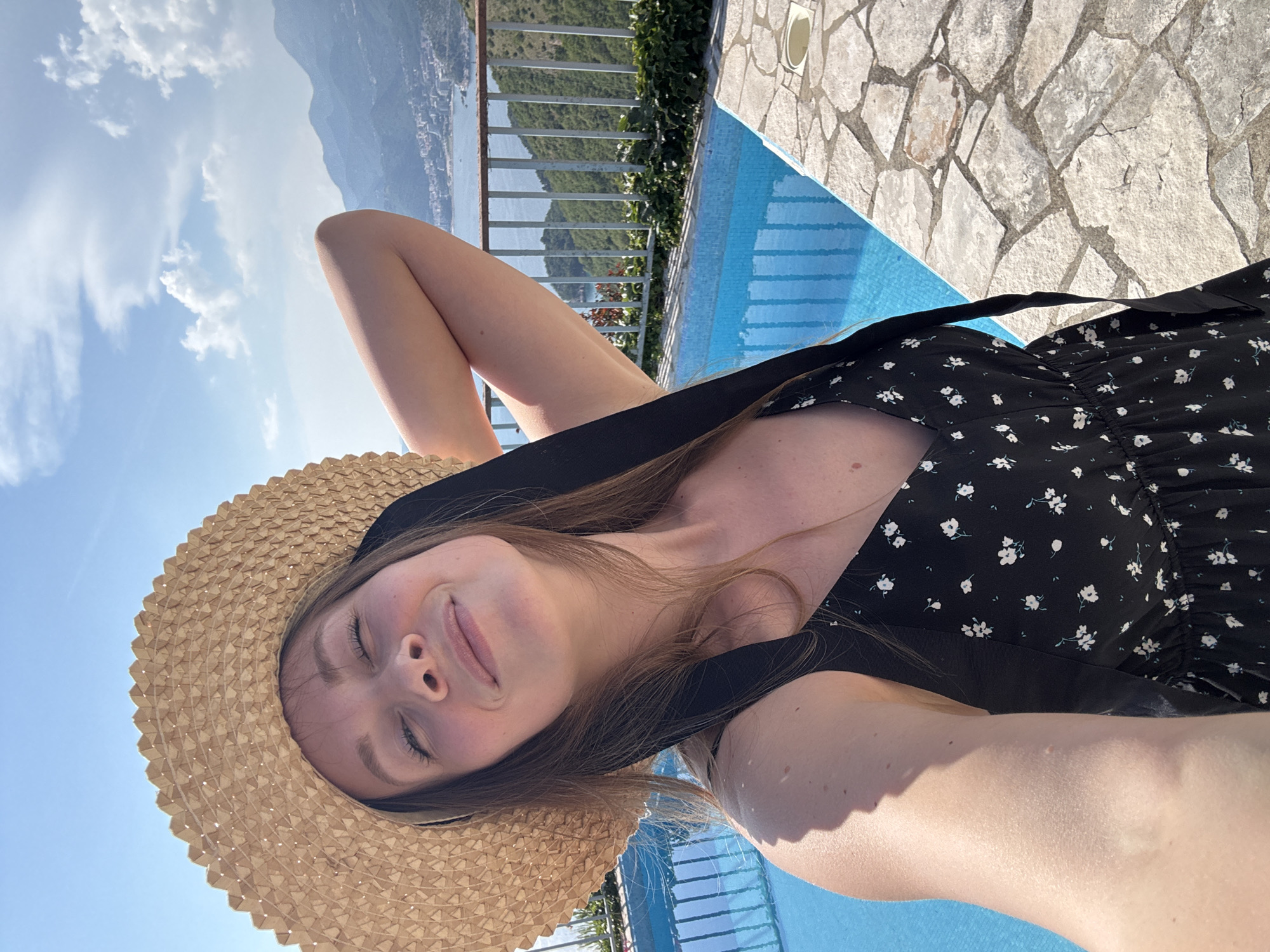 In Sveti Stefan, Montenegro, a woman wearing a straw hat and a black floral dress takes a selfie outdoors. She is positioned near a bright blue swimming pool, with a paved area and a metal railing in the foreground. Beyond the railing, lush green hills and distant mountains are visible, leading down to a body of water and a coastal town. The sky above is a clear blue with scattered white clouds. The woman's eyes are closed, and she has a relaxed expression on her face as she holds her right arm up.