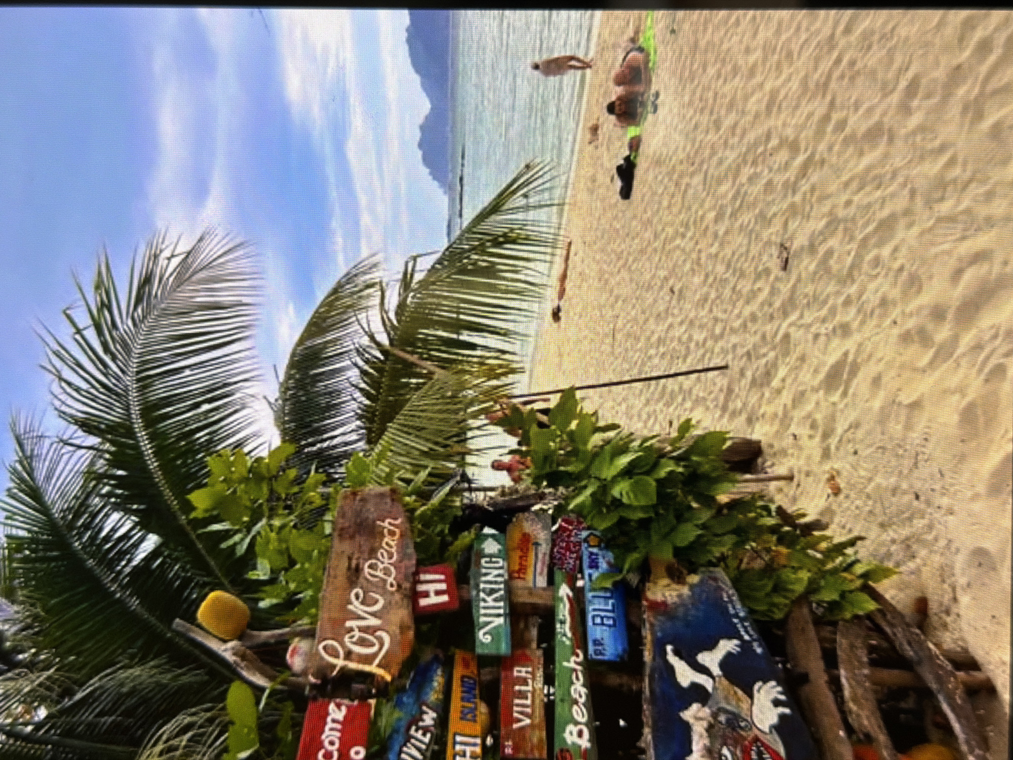 The image depicts a vibrant and sunny tropical beach scene, oriented vertically. In the immediate foreground on the left, large green palm fronds extend into the frame, partially obscuring a clear blue sky with soft white clouds. Below the palms, a dense arrangement of green foliage surrounds a collection of rustic, hand-painted wooden signs.
These signs, varying in color and shape, serve as direction markers. Prominently visible text on these signs includes "Love Beach" on a weathered brown plank, along with "HI", "Viking", "Villa", "Beach", "PP. BLU", "Come", "View", and "HI Island". A single yellow fruit, resembling a lemon or lime, is nestled within the greenery near the signs.
Beyond the foliage and signs, the scene opens to a wide expanse of light golden sand, showing gentle undulations. Several people are enjoying the beach: one individual is standing in the shallow, clear turquoise water, while another is relaxing on a green mat on the sand, seemingly sunbathing. Other figures are less distinctly visible further along the shore.
The tranquil ocean stretches into the background, with its calm surface reflecting the bright sky. In the distance, large, hazy blue mountains or islands rise majestically from the water. The sky above is a bright, clear blue with scattered white clouds, suggesting a beautiful, sunny day. The overall atmosphere is one of relaxation and tropical leisure. The location, based on visual cues, appears to be a Southeast Asian or similar tropical beach, which contradicts the provided "Lovech, Bulgaria" context.