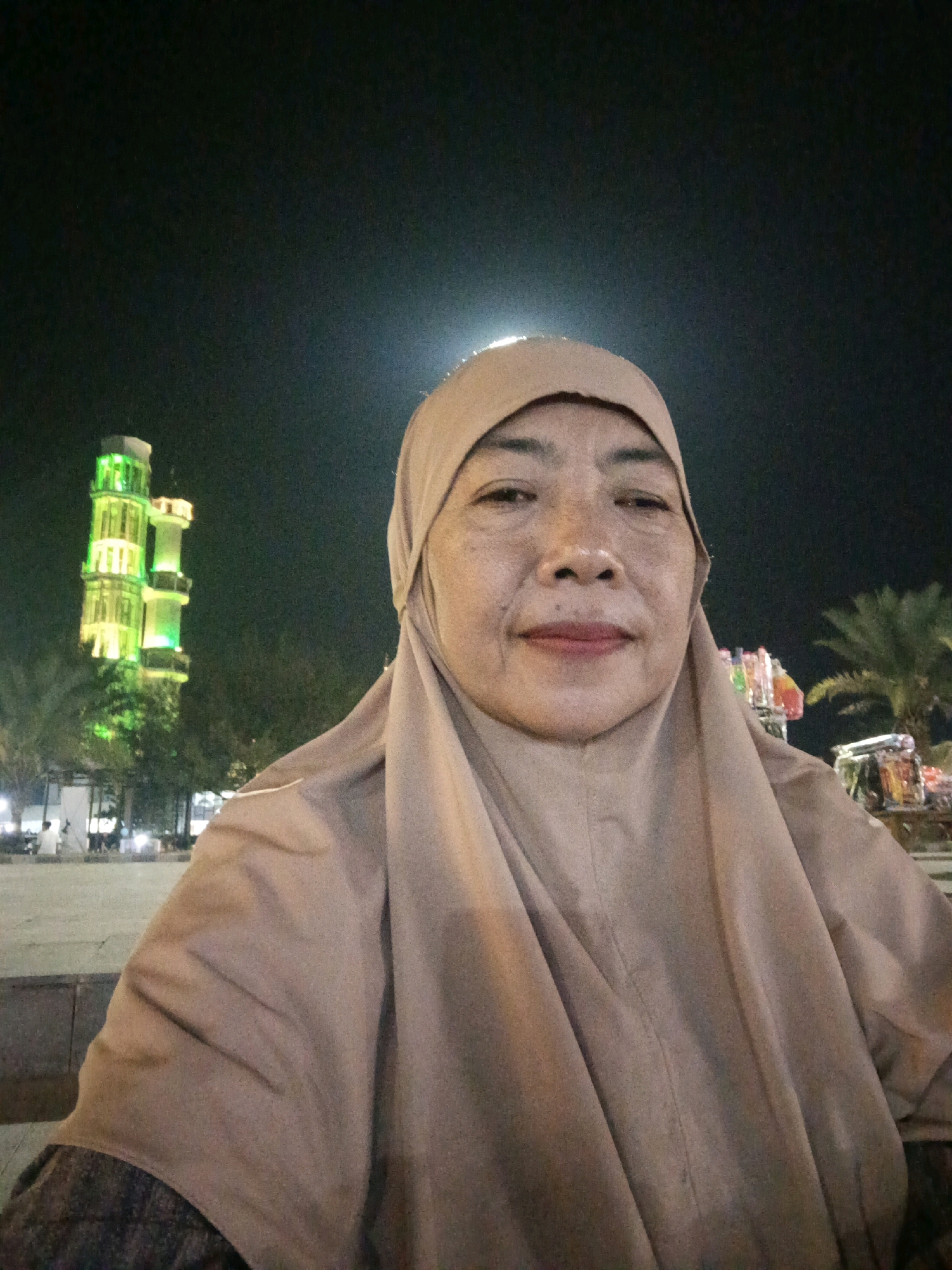 The image is a selfie taken at night in the city of Lubuklinggau, Indonesia. The subject is an adult woman wearing a light brown hijab, with her face prominently in the foreground. In the background, a tall, narrow tower-like structure with green lights is visible, along with palm trees and other buildings. The lighting suggests it is nighttime, with artificial lights illuminating the scene.