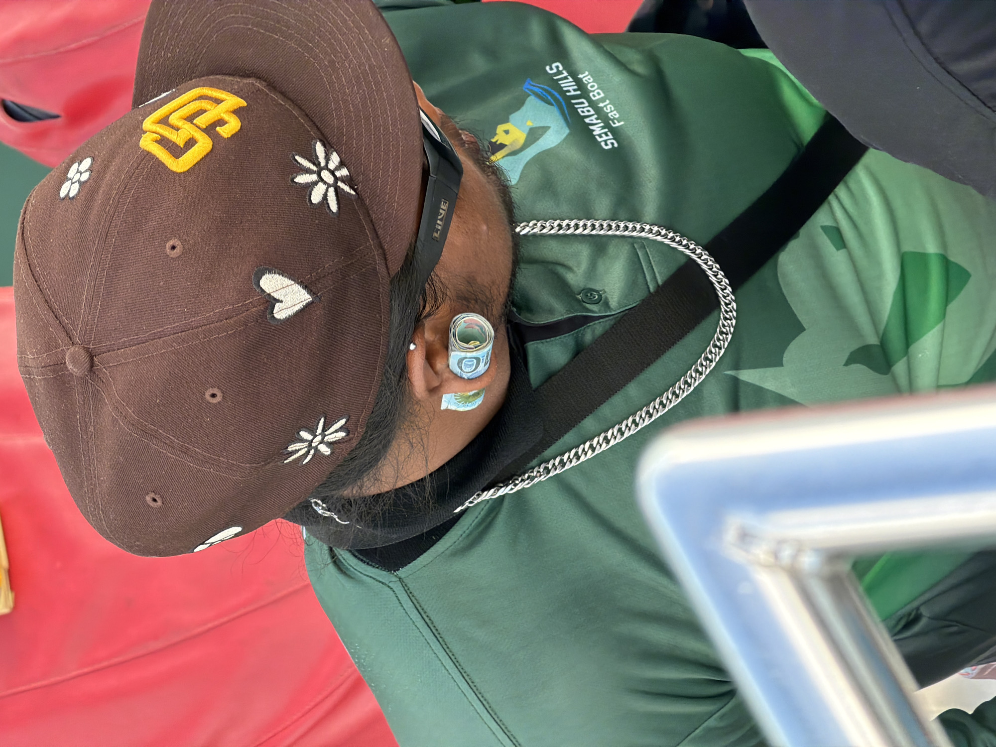 The image shows a close-up, angled view of a person, likely male, from the neck up, adorned with a baseball cap and a silver chain. The person is wearing a dark green shirt or jacket with the white text "SEMABU HILLS Fast Boat" and a distinctive logo featuring a yellow figure in a boat. This text strongly indicates the setting is a fast boat service, consistent with operations in Bali, Indonesia, given the provided location context of Denpasar.
The brown baseball cap prominently displays a yellow "SD" logo, along with embroidered white flowers and a white heart design. A striking detail is a rolled-up 10,000 Indonesian Rupiah banknote (indicated by the visible "10" and design) tucked into the person's right ear. A black strap, possibly from headphones or a bag, with the word "LINE" on it, is also visible near the ear.
The background includes a blurry red surface, likely a seat or deck, and an out-of-focus silver metal railing in the foreground, reinforcing the boat setting. The lighting suggests it is daytime, capturing a casual moment of travel.