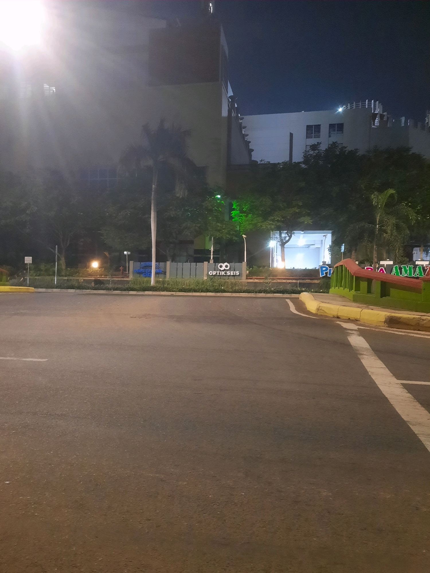 The image shows a nighttime scene in front of a building, likely in a commercial area. The foreground is dominated by a paved road with a white lane marking. To the right, a yellow curb separates the road from a green and red painted structure that appears to be a decorative barrier or railing. Beyond this, a portion of a building with illuminated text, possibly a business name, is visible. The text includes "P" and "AMA". To the left, a sign with a logo resembling two circles and the text "OPTIK SEIS" is present. In the mid-ground, there are trees and palm trees, some illuminated by artificial lights. A bright light source, possibly a street lamp or building light, is visible in the distance, creating a hazy glow. In the background, a multi-story building with windows is dimly lit. The overall impression is a well-lit urban environment at night. While the image does not explicitly state its location, the presence of the business "Optik Seis" suggests a commercial setting.