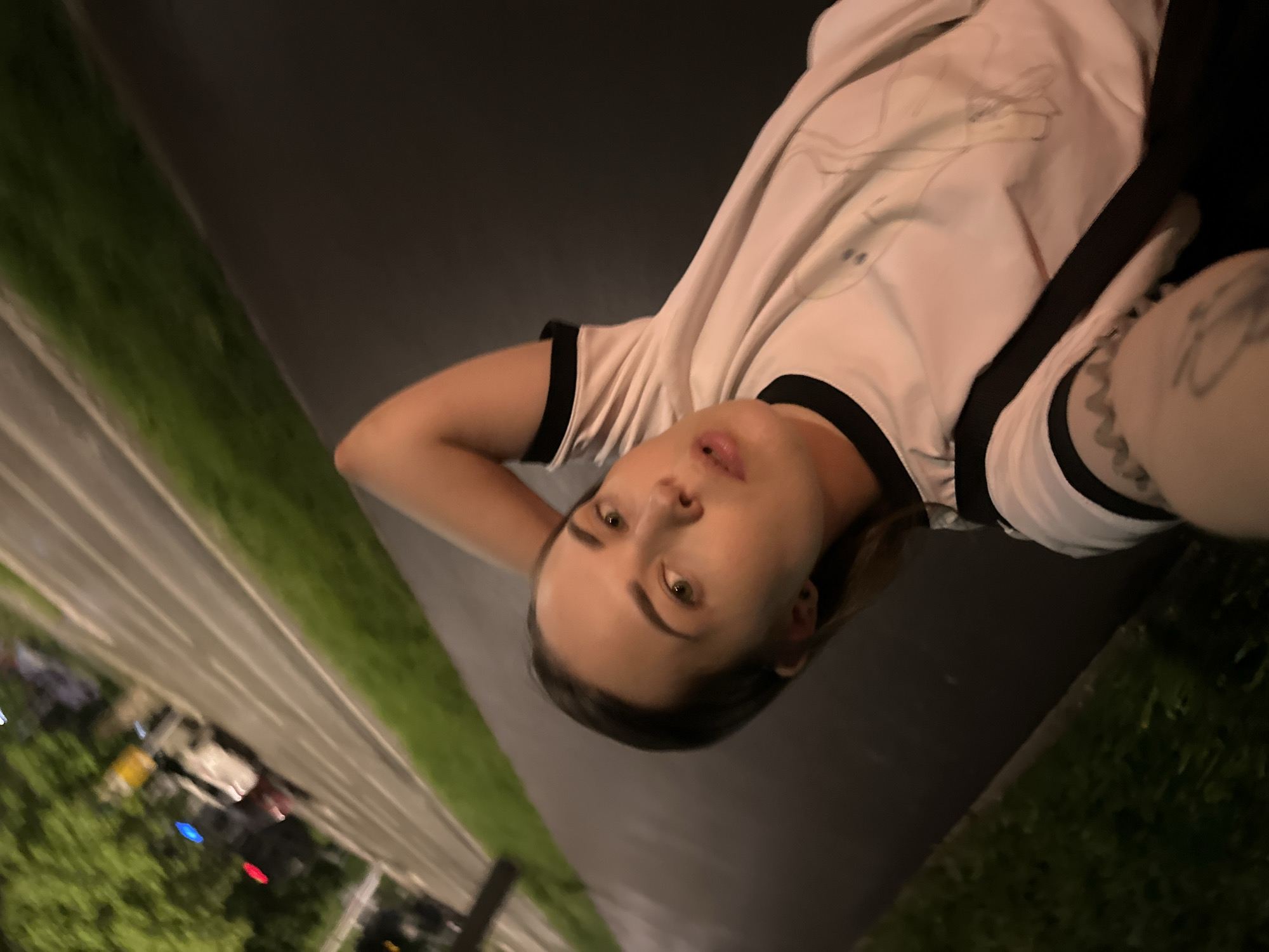 A young adult woman is lying on the edge of a sidewalk at night, holding her camera at arm’s length to take a selfie. She has light skin, green eyes, and dark hair pulled back from her face. She is wearing a white t-shirt with black trim around the collar and sleeves. Part of a tattoo is visible on her arm. The sidewalk is dark gray and appears to be adjacent to a grassy area with trees. Beyond that is a multi-lane road, with cars driving on it. Streetlights are visible and the lights of the cars are on. Based on the visible environment and details, it seems to be the city of Perm, Russia.