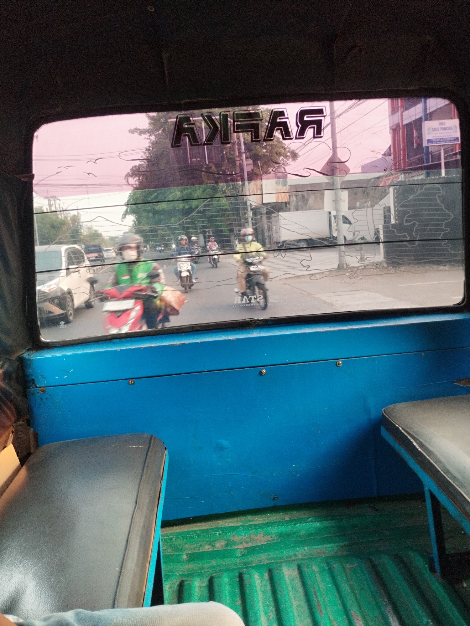 The image is taken from inside a vehicle, looking out through a pink-tinted rear window. The window has a dark graphic decal on it that reads "RAFAAR". Outside the vehicle, a street scene unfolds in what appears to be Surabaya, Indonesia.
Several people on motorcycles are visible. One person in a green jacket and helmet is riding a red and white motorcycle. Another person in a yellow jacket and helmet is on a different motorcycle, with "RATZ" visible on the side of it. Further down the road, other motorcycles and vehicles can be seen. Buildings line the street, with some signage visible. The sky is overcast, and trees are present.
Inside the vehicle, the blue interior of the passenger compartment is visible, with a green metal floor and two dark-colored seats. One seat on the right is partially visible, and a portion of another seat on the left is also in view. The image is grainy and appears to have been taken with a mobile phone.