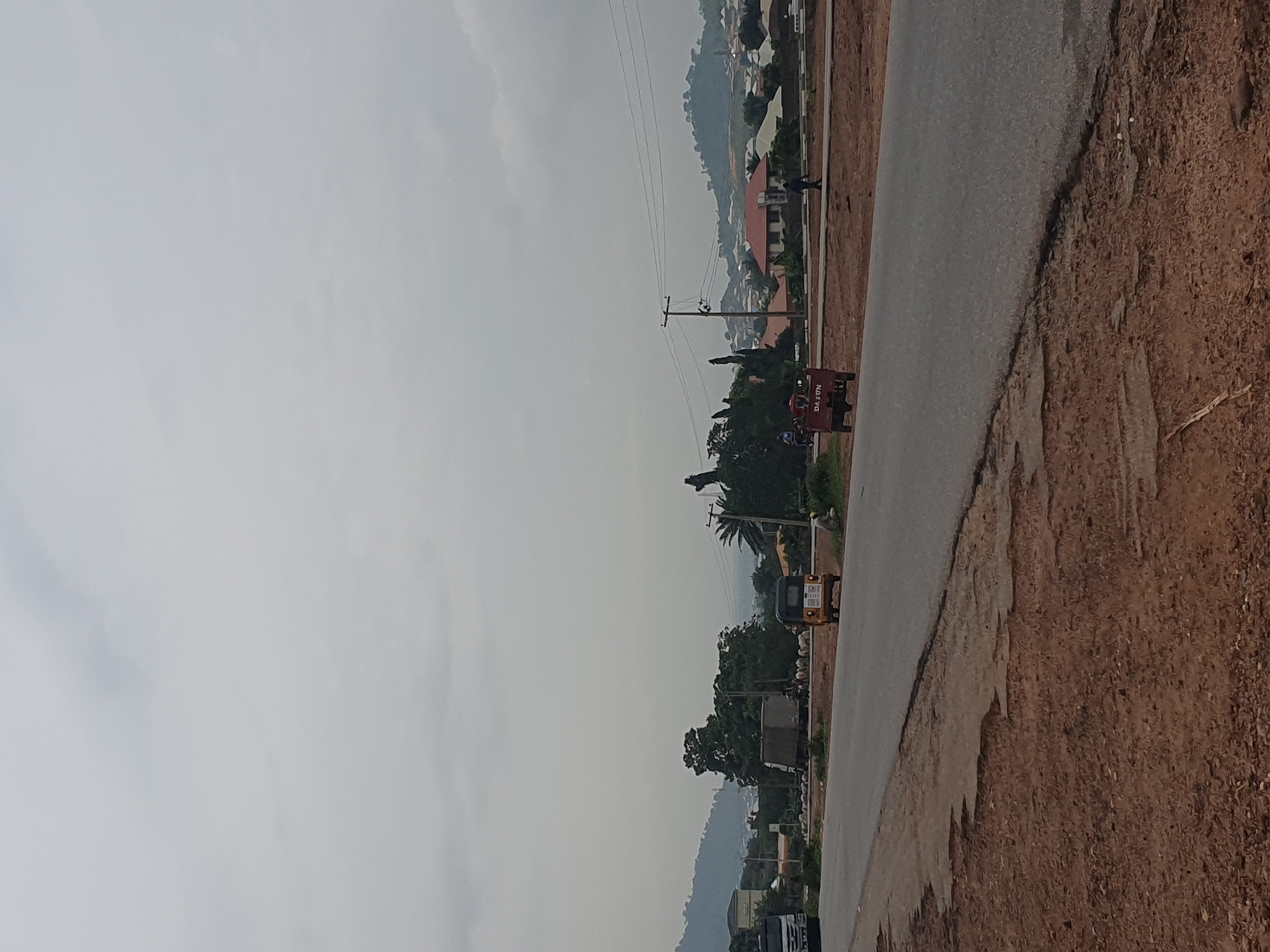 The image depicts a roadside scene in Lokoja, Nigeria, under an overcast sky. In the foreground, a paved road with a broken edge runs diagonally from the bottom left to the top right. The roadside verge is composed of dry, reddish-brown soil and gravel.
Further down the road, several vehicles are visible. A white truck is parked on the left side of the road. To its right, a herd of sheep is gathered near a stationary truck with a grey container. A yellow auto-rickshaw is also parked on the side of the road. In the middle distance, a red motorized tricycle is traveling on the road.
In the background, a line of trees separates the road from a developed area with houses featuring red and white roofs. Beyond the houses, rolling hills covered in vegetation rise up, with some buildings scattered on their slopes. Utility poles with overhead wires are present along the roadside. A few individuals are visible walking or standing near the vehicles and houses. The overall impression is of a semi-urban or peri-urban environment.