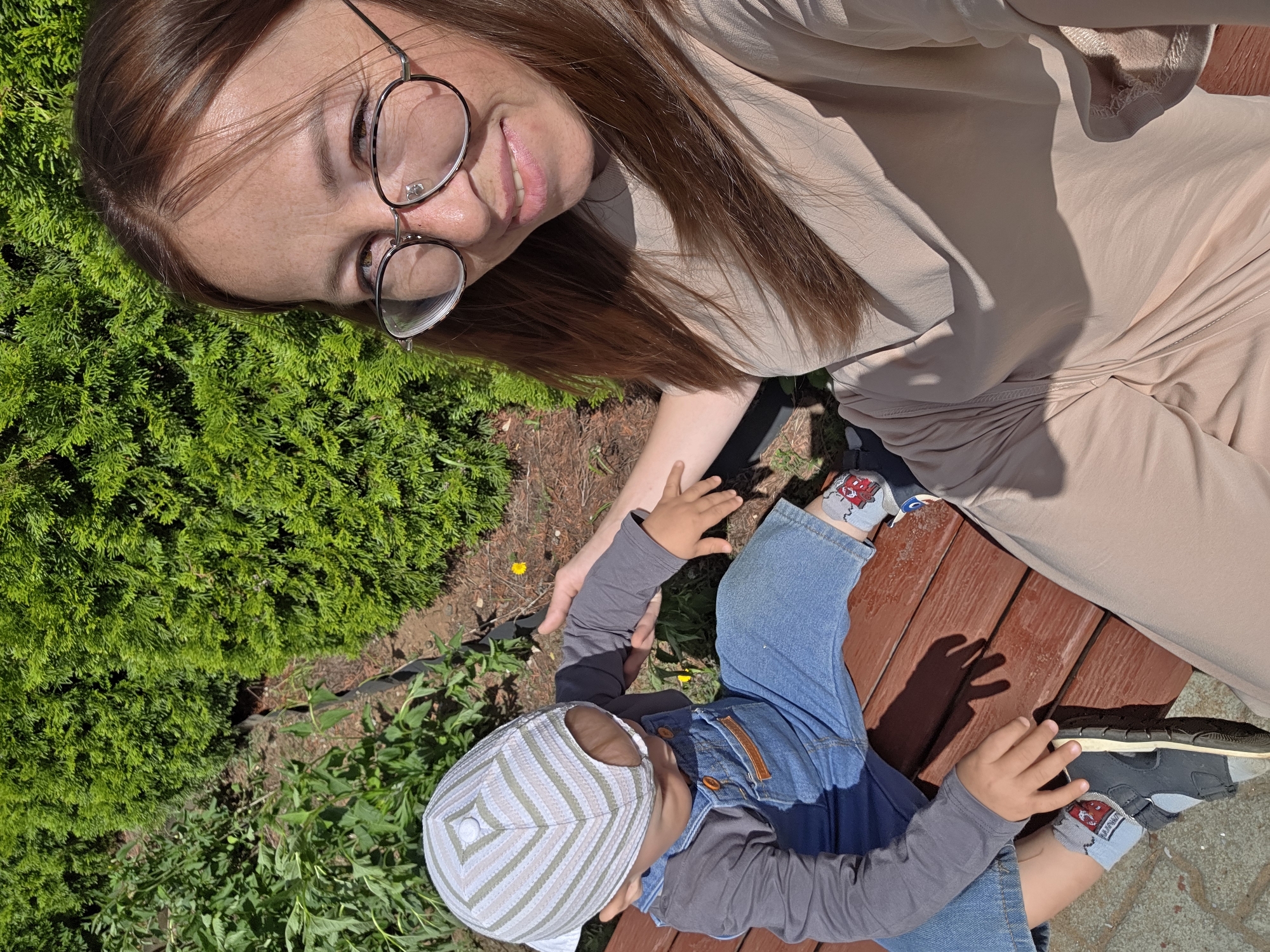 An outdoor, high-angle selfie captures a woman with brown hair and glasses, smiling at the camera, seated on a wooden bench next to a young child wearing a cap and denim overalls. The background consists of lush green foliage and a patch of sunlight. The woman is wearing a beige t-shirt and pants. The child is wearing a gray long-sleeved shirt and blue denim overalls, with a striped cap and grey sneakers. The setting appears to be a park or garden area. The presence of Thuja trees and deciduous plants suggests a temperate climate, consistent with Chaikovsky, Russia. The lighting indicates it is daytime, likely late morning or early afternoon given the angle of the shadows.