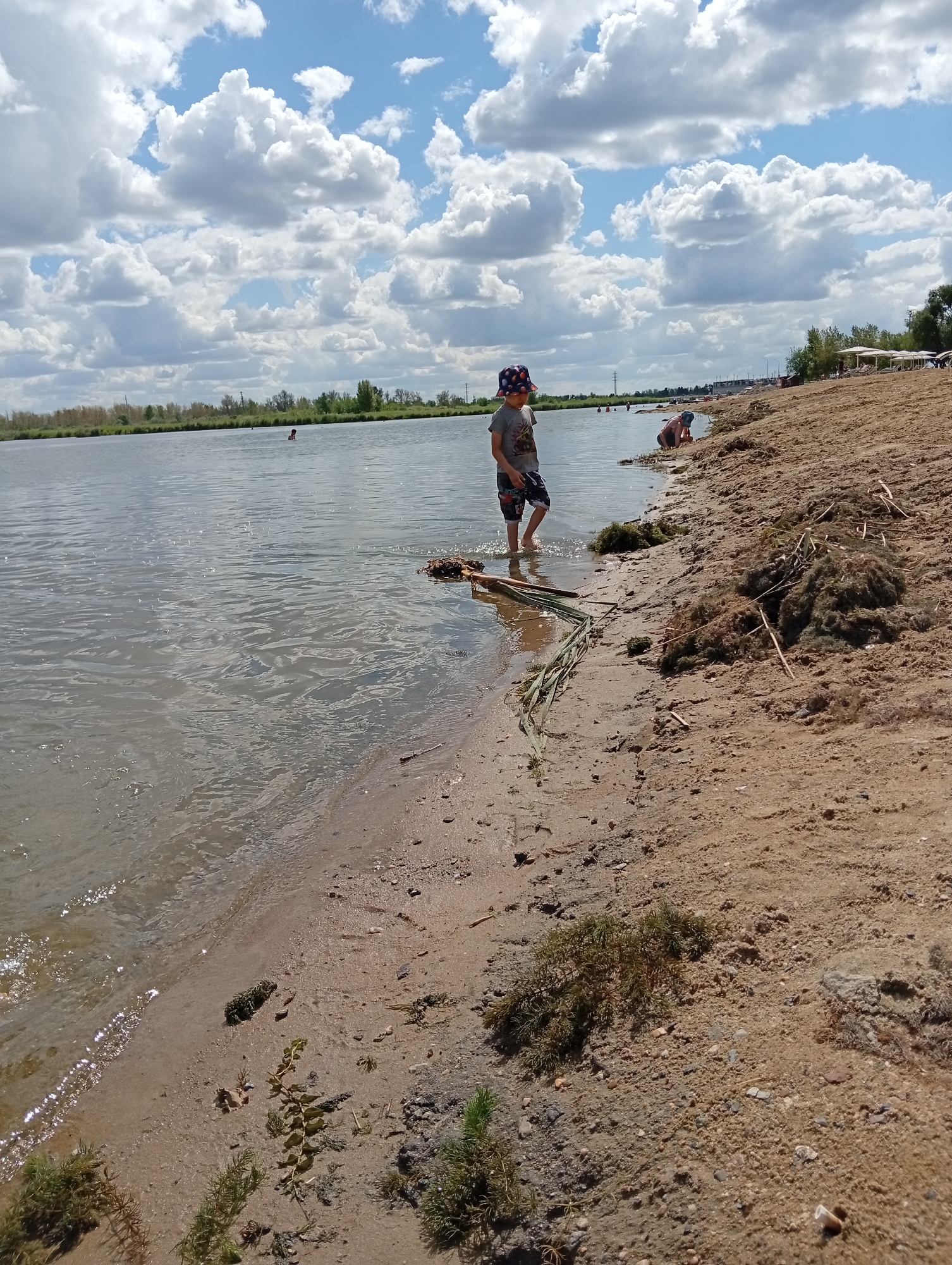 The image depicts a sunny day at a beach, likely within the Мичурин ауылдық округі, Kazakhstan. The foreground is dominated by a sandy shore with scattered vegetation and debris. In the middle ground, a young boy wearing a hat and shorts stands in shallow water, facing away from the camera. Further in the water, other figures can be seen, possibly swimming or wading. The background shows a tree line along the water's edge and a cloudy sky. Several white beach umbrellas are visible in the distance on the shore, suggesting a recreational area. The water appears calm with some rippling.
