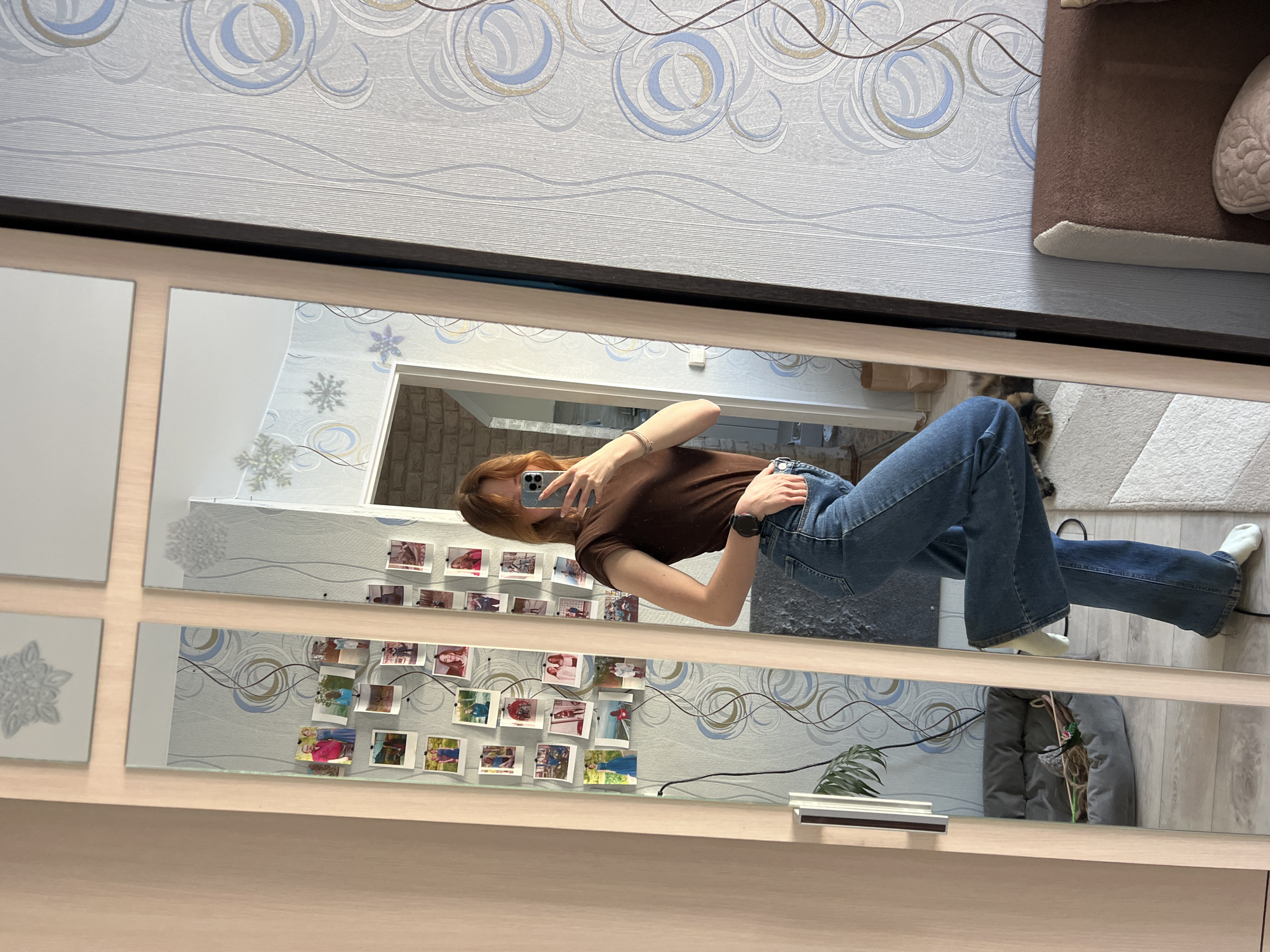 A young woman with reddish-brown hair is reflected in a large mirror, taking a selfie with her smartphone. She is wearing a brown t-shirt and blue jeans. Her left wrist has a bracelet, and she is wearing a black watch on her left wrist. The mirror reflects a room with patterned wallpaper, multiple photos displayed on the wall, and parts of the room including a cat bed, a gray jacket or bag, and a potted plant. Part of the cat can also be seen in the mirror. The location is possibly in an interior setting in Chaikovsky, Russia, based on the style of the interior decoration.