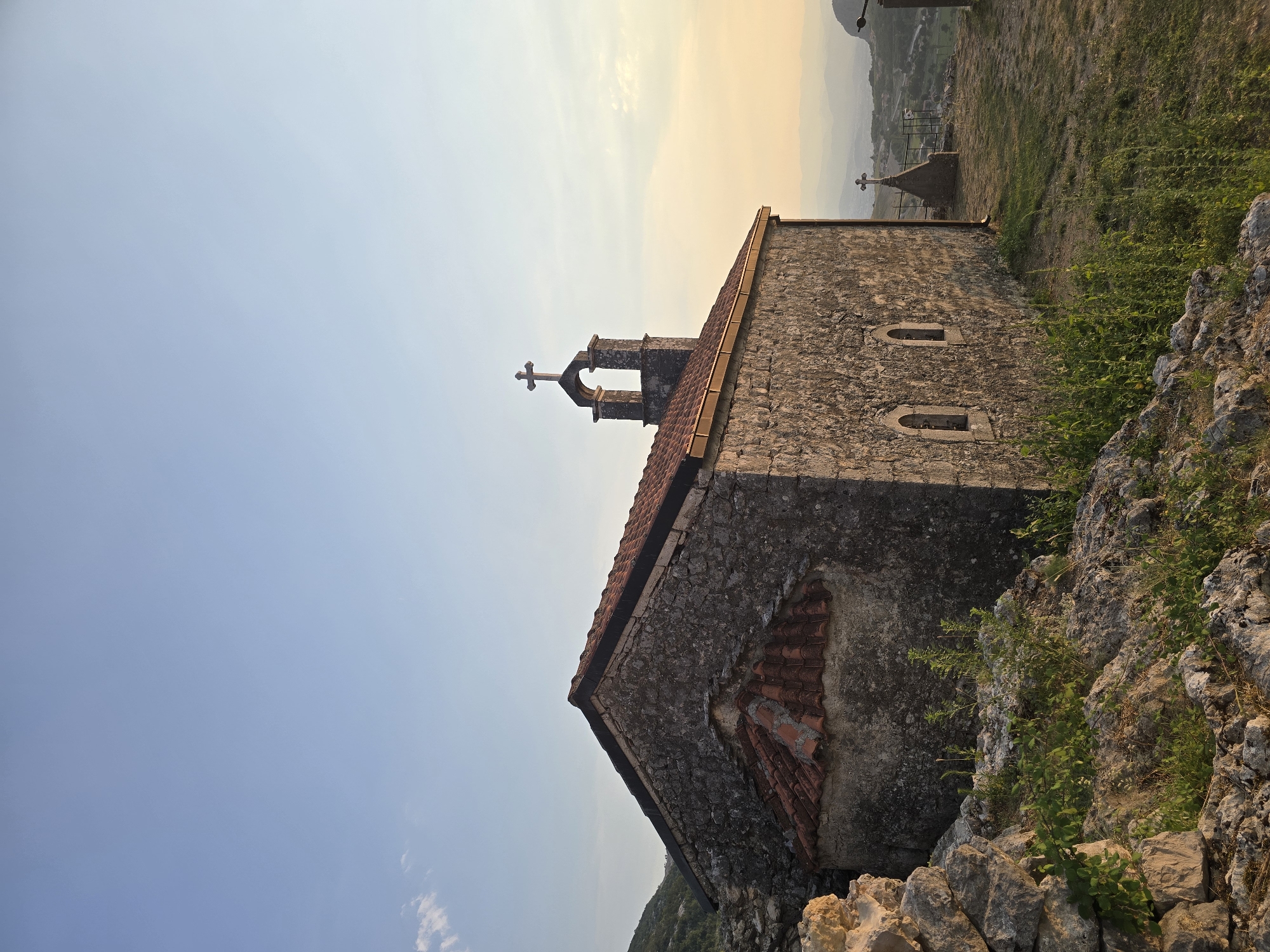 The image shows a small, stone-built chapel situated on a hilltop in Medun, Montenegro. The chapel has a red-tiled roof and a small bell tower topped with a cross. Two arched windows are visible on the side of the chapel. In the foreground, there are rocks and green vegetation. In the background, rolling hills and a distant town can be seen under a pale, hazy sky. Another cross is visible on a pedestal to the right of the chapel. The overall scene evokes a sense of peace and historical significance.