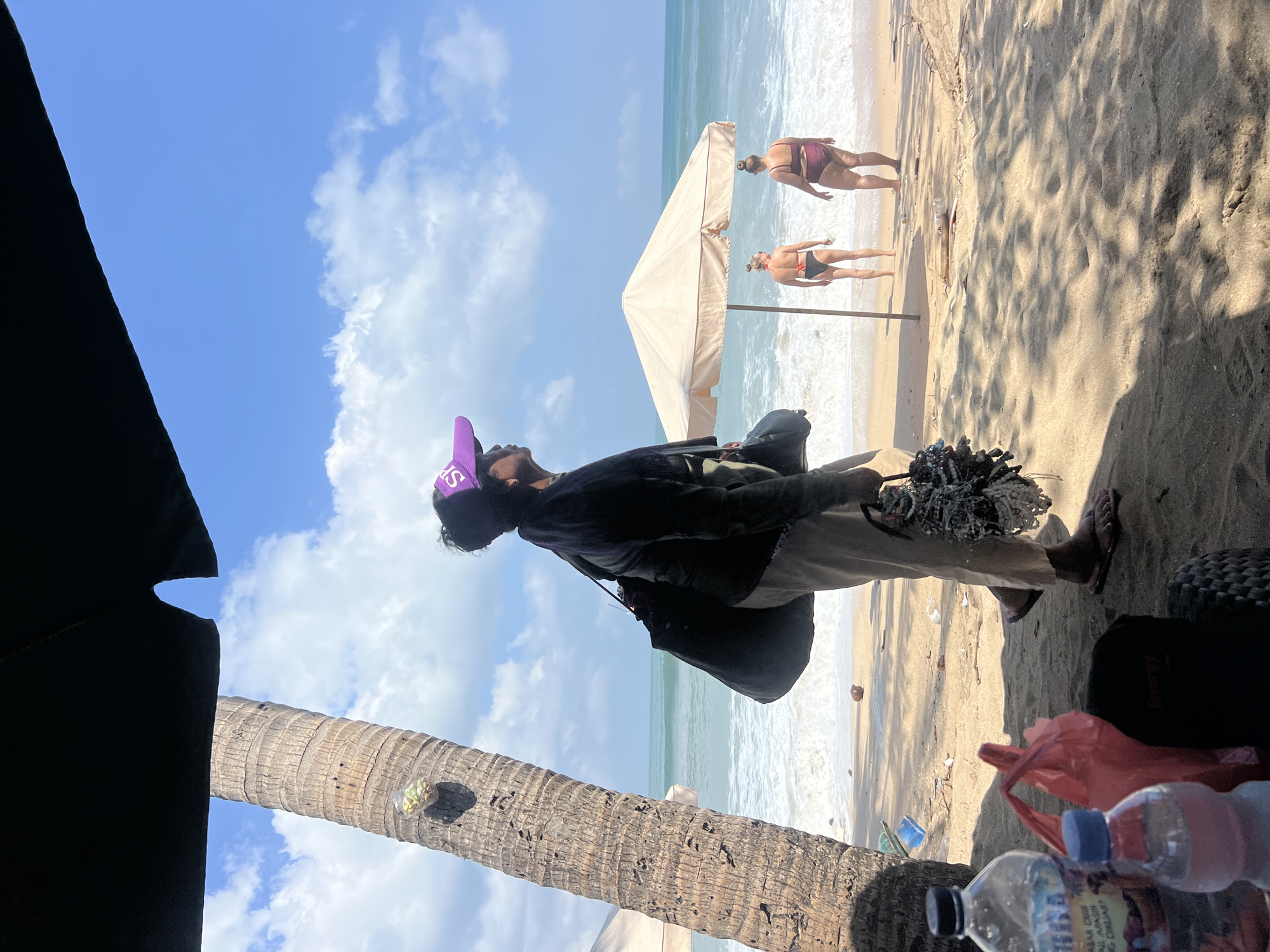 A person wearing a purple visor and a dark top stands on a sandy beach in Kuta, Indonesia. They are carrying a large black bag over their shoulder and a bundle of decorative items in their right hand. In the background, two individuals in bikinis stand near a beige umbrella at the edge of the water, with waves breaking on the shore. The sky is blue with scattered white clouds, and a palm tree trunk is visible on the left side of the frame. Bottles of water and red plastic bags are placed on the sand in the foreground.