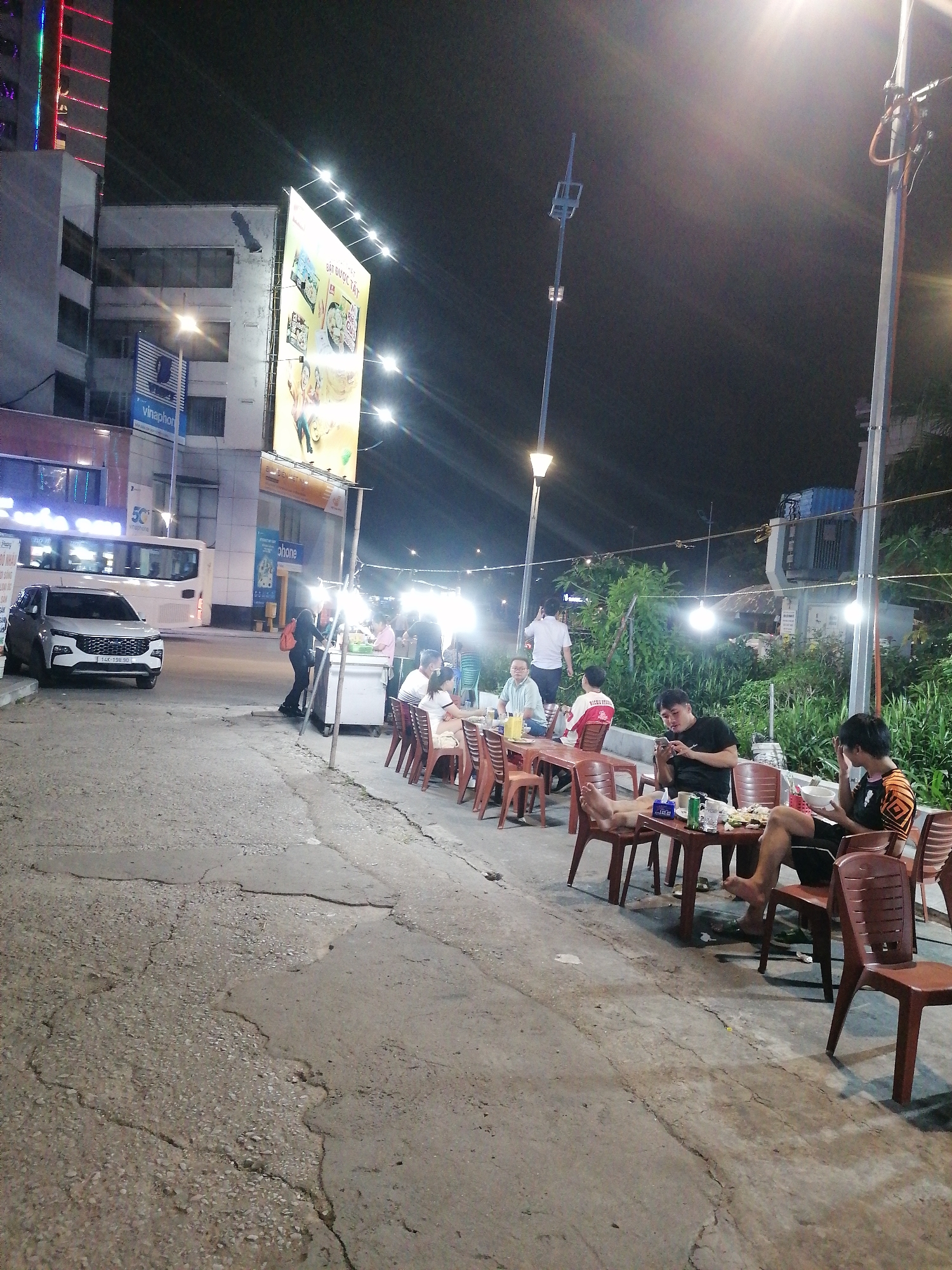 People are eating at tables set up on the side of a street in Bai Chay Ward, Vietnam, at night. Several tables with brown plastic chairs are occupied by individuals eating and socializing. The street is cracked and uneven. A food cart is visible near a bus. A large billboard is illuminated above buildings on the left. Streetlights and string lights provide illumination. Lush green vegetation is present behind the diners.