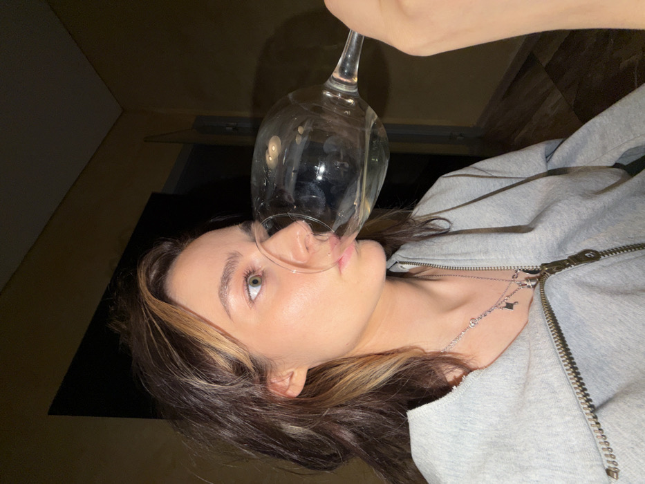 A person wearing a gray zip-up hoodie holds a large wine glass to their face, the rim pressing against their nose and upper lip. The person's face is visible, revealing light skin, light green eyes, and brown hair with blonde highlights. A silver necklace adorns their neck. The background consists of dark-toned walls and a partially visible wooden floor. The image is taken at an angled perspective.