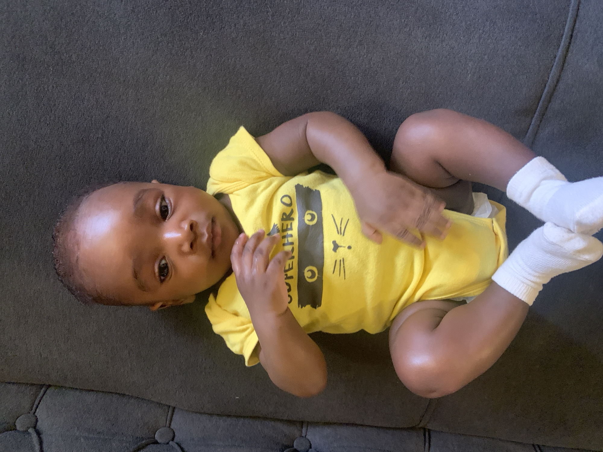 A baby lies on a gray tufted couch. The baby wears a yellow onesie with a black cat-like superhero emblem on the front and white socks on the feet. The location in the city of Barakin Akawo, Nigeria, is not discernible from this image.