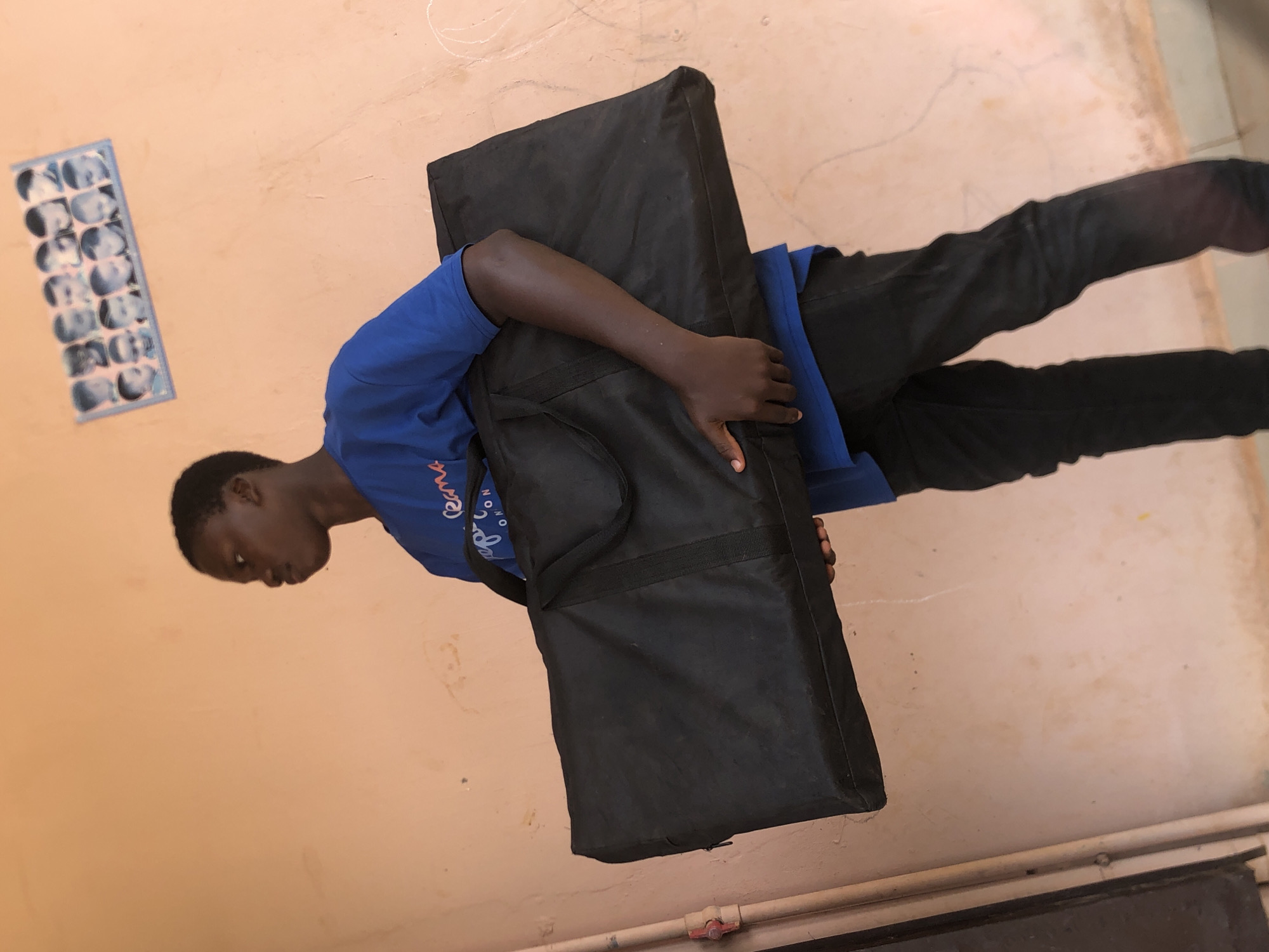 An African male stands indoors in Nkwelle Ezunaka, Nigeria. He's carrying a black, rectangular bag across his body. He wears a blue t-shirt and black pants. The background is a light beige wall. A rectangular picture frame with multiple small headshots is attached to the wall.
