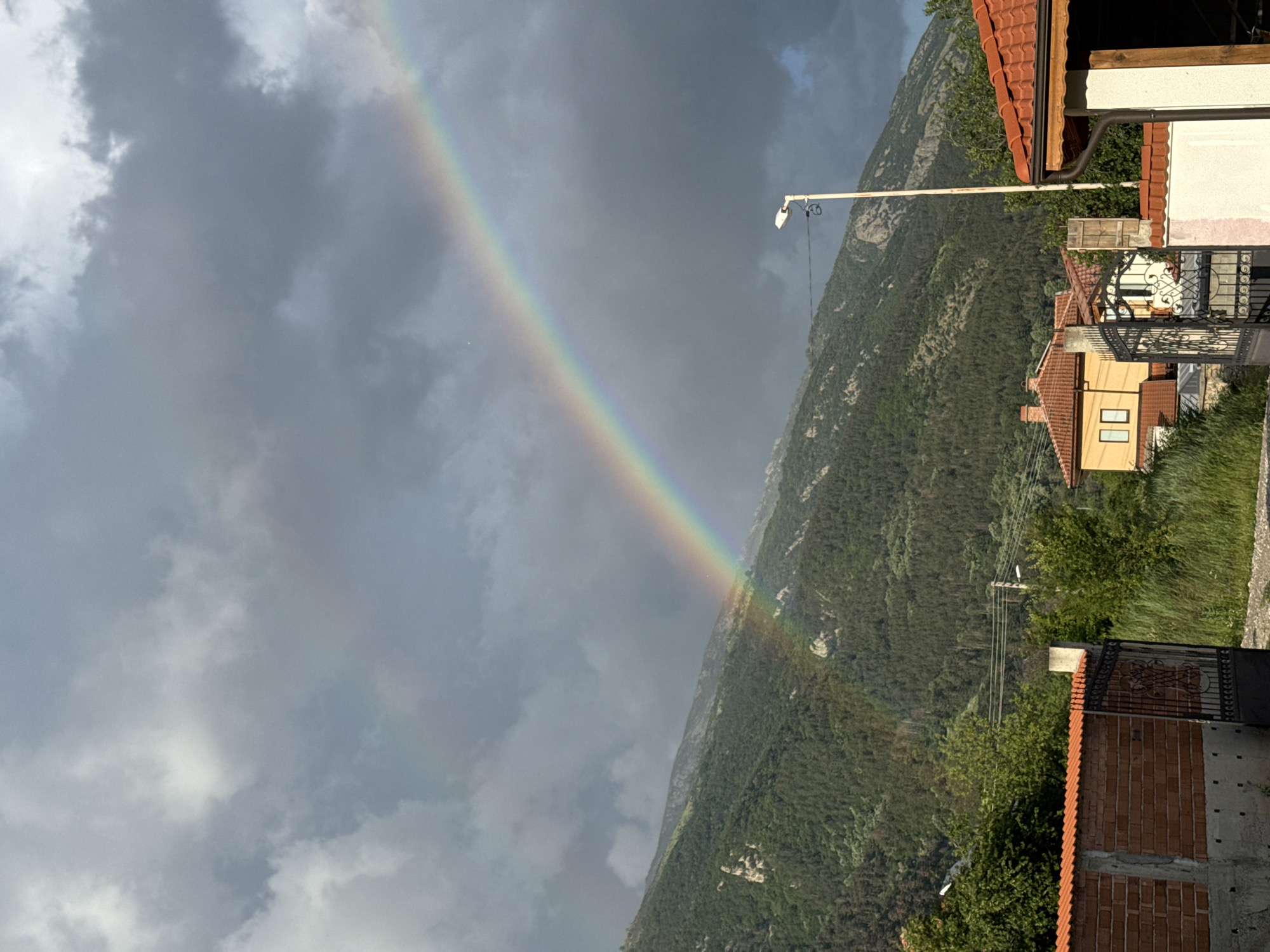 The image shows a rainbow ending near a mountain covered in trees, with houses on the right side of the frame. The sky is cloudy. The location is in Unknown, Unknown.