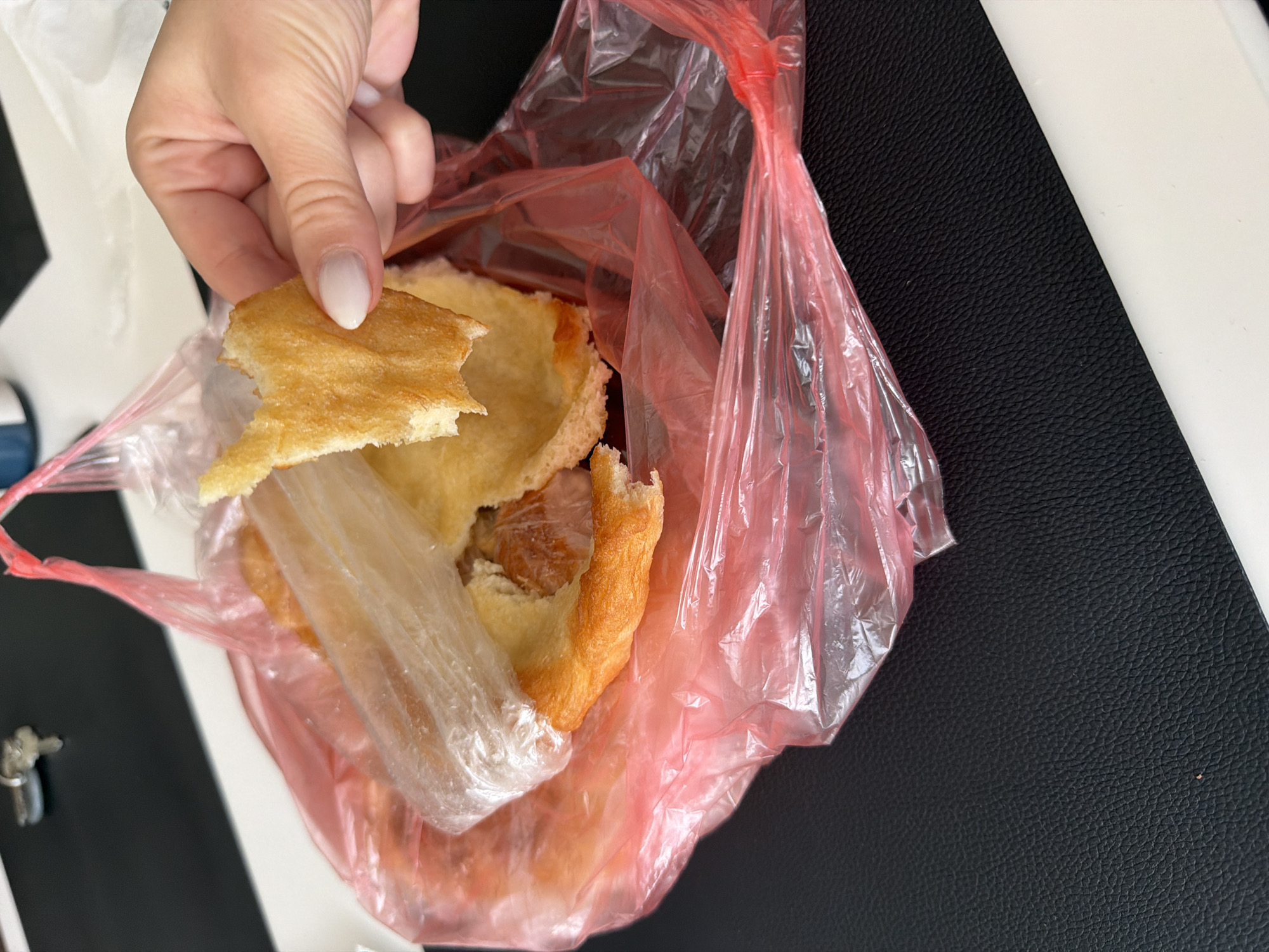 A hand holds a piece of fried dough over a red plastic bag containing similar fried dough, likely mekitsa, in what is probably Plovdiv, Bulgaria. The fried dough appears golden brown with an irregular edge where a bite has been taken. The red bag sits on a surface that is partially white and partially black. A set of keys is visible in the lower left corner.