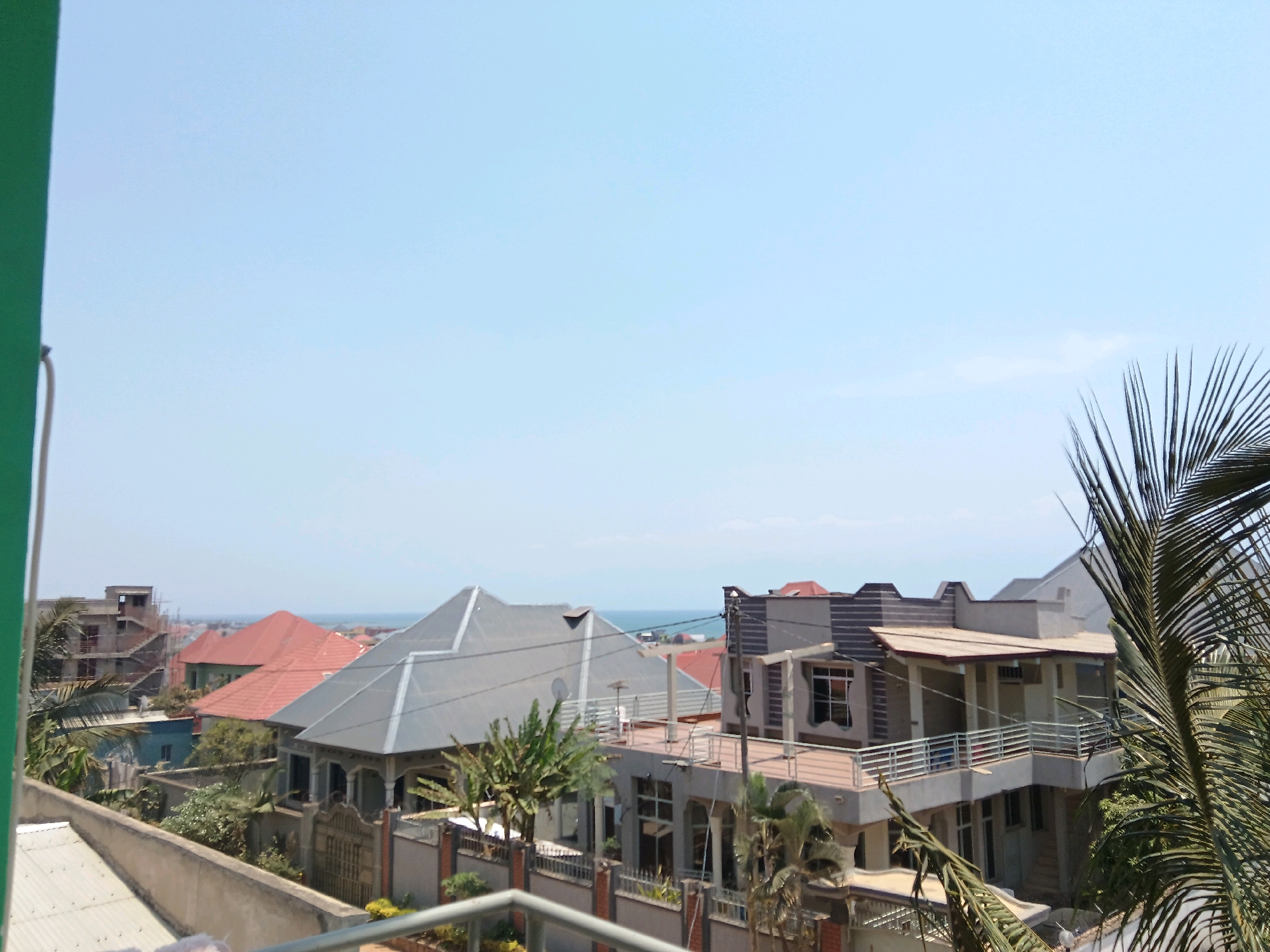 The image displays a view of houses and buildings in Gitaramuka, Burundi, with the ocean visible in the distance. The scene is bathed in daylight under a clear sky. In the foreground, on the left, a portion of a green structure is visible, possibly a balcony railing or part of a building. The middle ground features several residential structures with distinct architectural styles. One prominent building has a grey, angular roof and multiple windows. To its left, another house has a red, pitched roof. Further back, a building under construction shows exposed concrete and scaffolding. Palm trees and other lush green vegetation are interspersed among the buildings. In the far distance, a thin line of blue suggests the presence of the ocean or a large body of water. The overall impression is of a moderately developed coastal area in an urban or semi-urban setting. There are no people visible in the image.