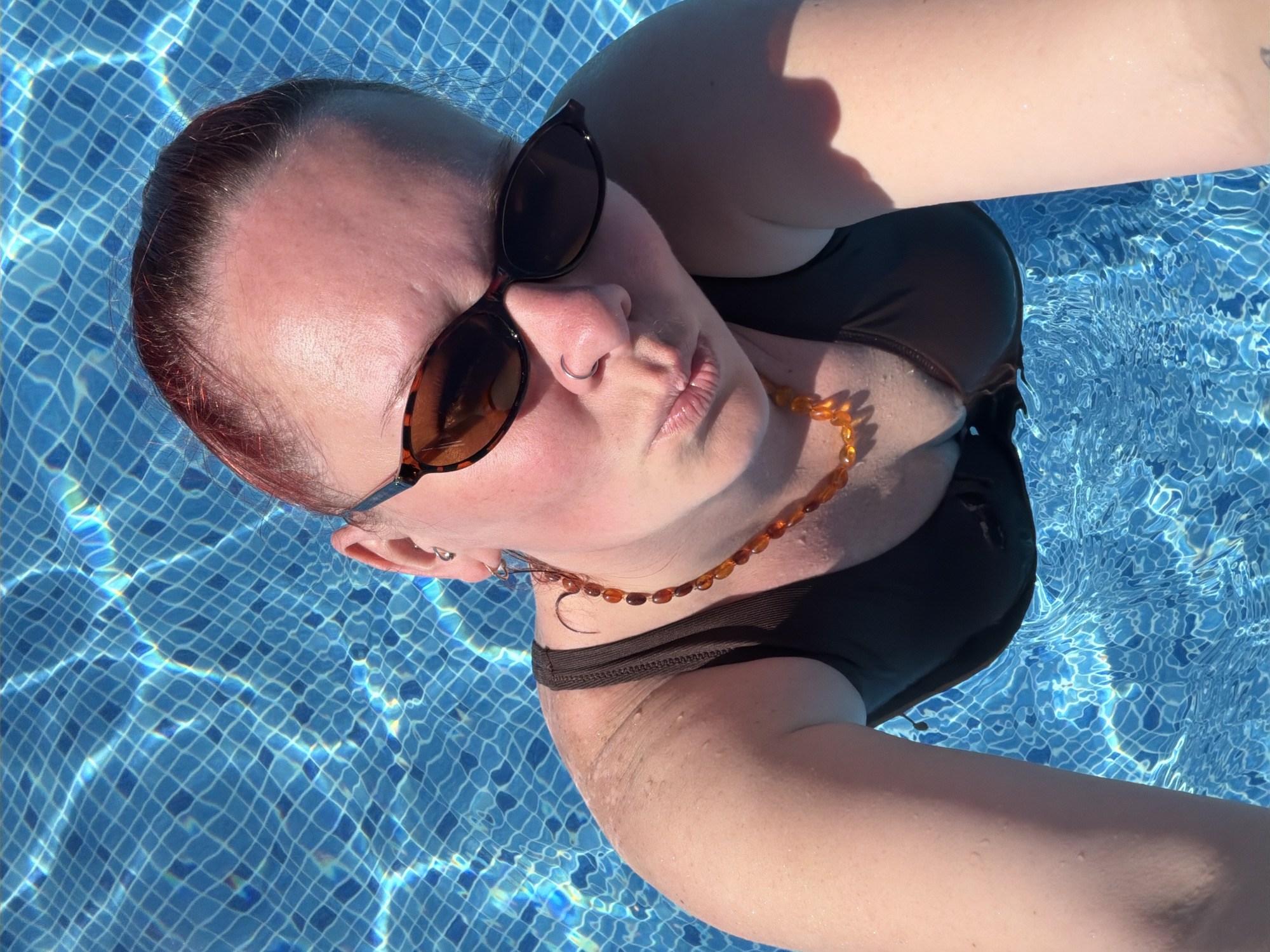 A person wearing sunglasses and a brown swimsuit is floating in a blue-tiled swimming pool. They have auburn hair pulled back and are wearing an amber-colored necklace and a nose ring. The pool's tile pattern is visible beneath the water. The image appears to be a selfie taken by the person in the pool. The location is not specified, therefore there is no way to determine if the photograph was taken in Votkinsk, Russia.