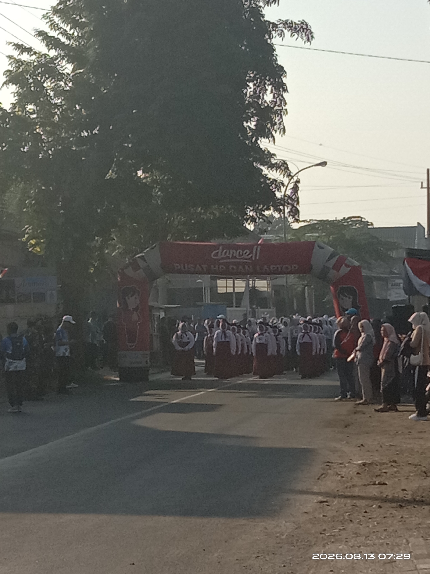 In Blimbing, Indonesia, an outdoor event is underway. A group of people, many wearing maroon skirts and white tops, stand in formation beneath a red inflatable archway. The archway is branded with the logo "dancell" and text that reads "PUSAT HP DAN LAPTOP". To the left of the arch, a few individuals, including one in a blue shirt and backpack, are visible on a paved road. The environment appears to be a street scene with trees on the left and buildings in the background. The time stamp indicates the date is August 13, 2026, at 07:29.