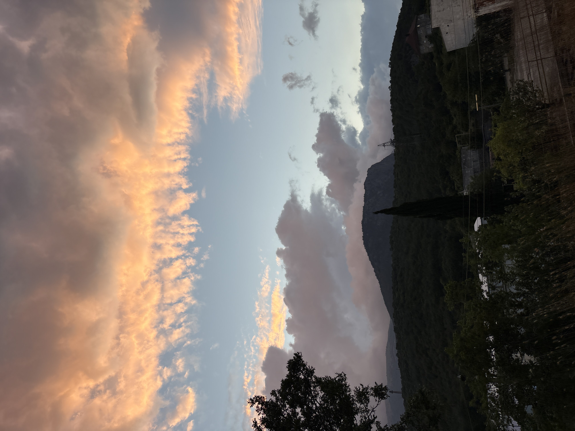 The image depicts a dramatic sunset over the town of Malyi Mayak, Ukraine. The sky is filled with an array of colorful clouds, ranging from deep oranges and pinks to soft blues and grays. These clouds are illuminated by the setting sun, creating a vibrant and dynamic display.
In the foreground, a dark silhouette of trees frames the scene on the left side. Below the sky and clouds, a lush green forested hillside stretches across the midground. Atop this hillside, a prominent mountain rises, its peak silhouetted against the colorful sky. A tall, slender cypress tree stands vertically on the slope, adding to the natural landscape.
Further down the hillside, signs of human habitation are visible. Buildings, including what appears to be a house with a dark roof and a white, somewhat dilapidated structure, are scattered among the trees. A power line tower with a lattice structure is also present on the hill. In the lower portion of the image, a more densely vegetated area with tall grasses and shrubs is visible, suggesting a natural or semi-wild area bordering the town. The overall impression is one of a natural landscape bathed in the dramatic light of a sunset, with human structures integrated into the environment.