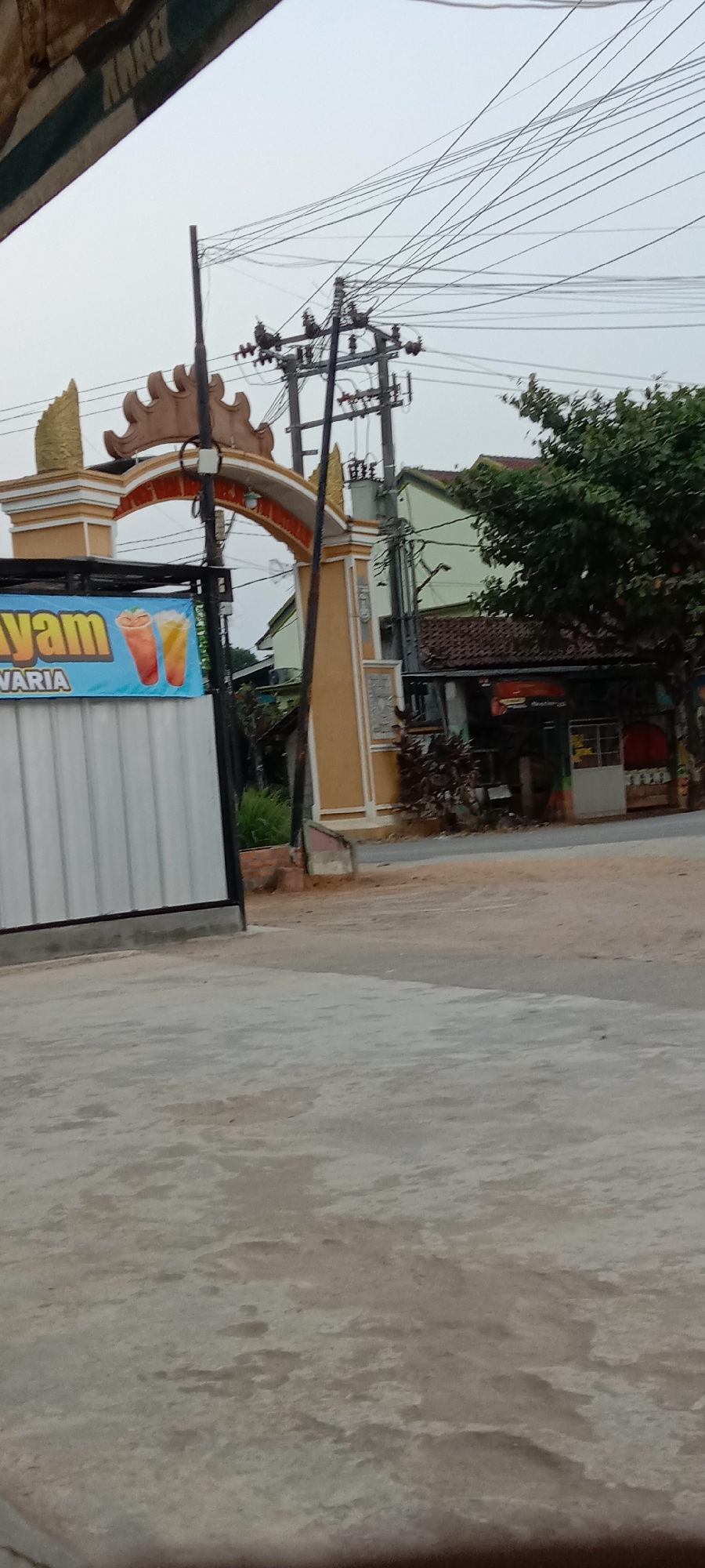 The image shows an arched gateway with decorative elements, likely an entrance to a complex or area in Utama Jaya Mataram, Indonesia. A banner is visible on the left, advertising "Ayam VARIA" with images of two colorful drinks, suggesting a food or beverage establishment. To the right of the gateway, there are buildings, including a structure with a red tiled roof and a green-painted upper story, and a smaller building with a white facade and a red awning. Utility poles and a dense network of electrical wires are prominent in the sky. The ground in the foreground is paved, appearing to be concrete, with some patches of dirt and gravel visible further back. Lush green trees are present on the right side of the image, obscuring some of the background. The overall scene suggests an outdoor commercial or residential area under an overcast sky.