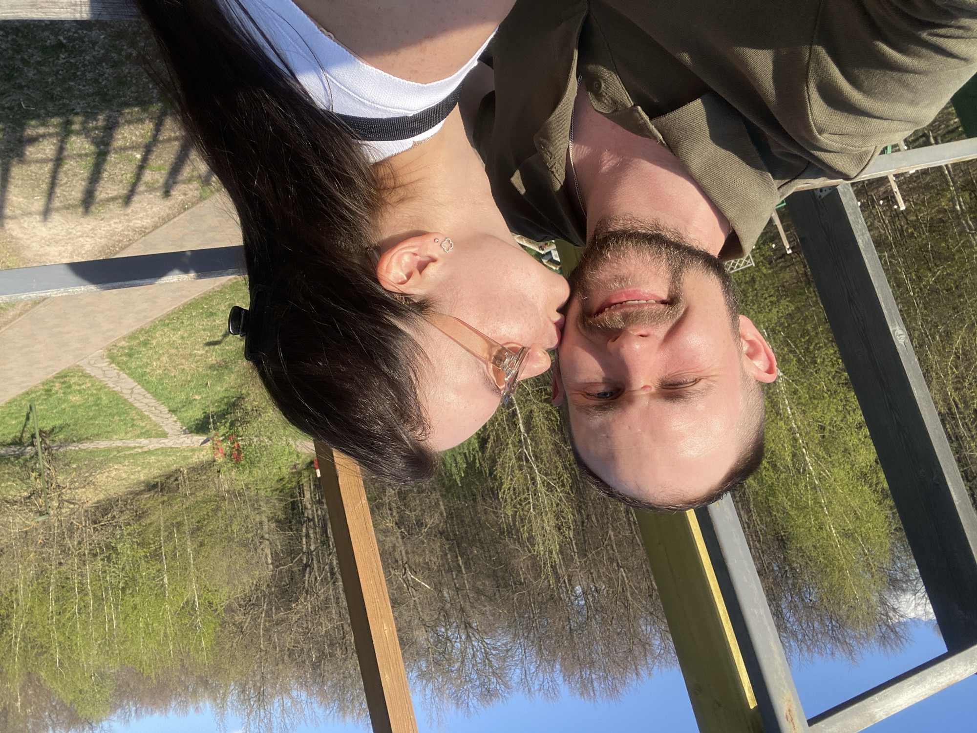 A man and a woman are posing for a photograph outdoors. The woman is kissing the man on the cheek. The woman has long, dark hair and is wearing glasses, a white tank top, and a black and white striped top underneath. The man has a beard and is wearing an olive green button-down shirt. They are standing on a wooden deck with a metal railing. In the background is a forest and a blue sky. The photograph was taken during the day. The exact location in Kazan, Russia, cannot be determined from the image.