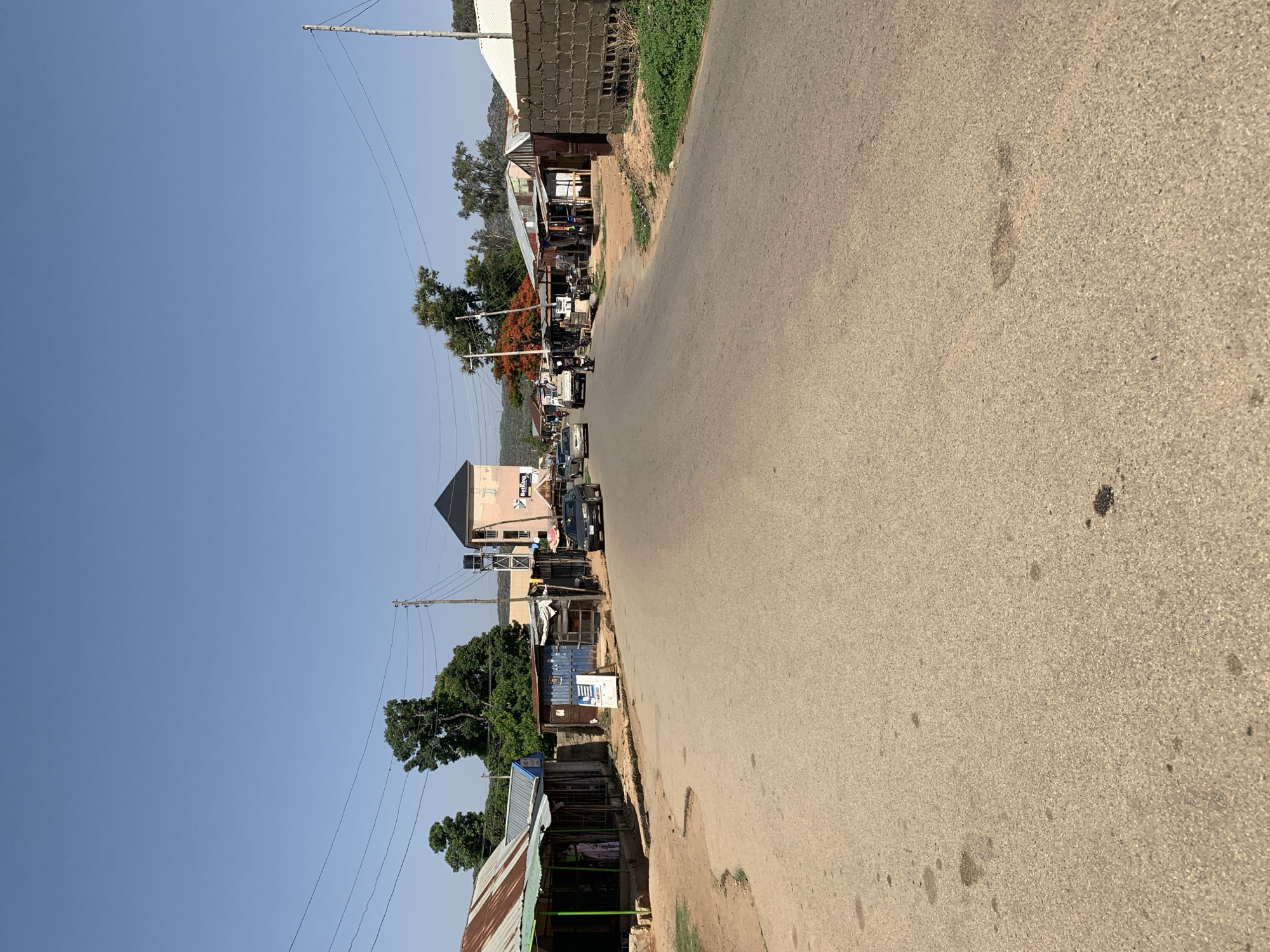 The image depicts a street in Rafin Par, Nigeria. The street is paved and shows signs of wear. Buildings of varying construction styles line the left side of the road, ranging from simple shacks with corrugated metal roofs to more substantial structures. A few vehicles are visible further down the street. Overhead power lines are present. The sky is clear and blue.