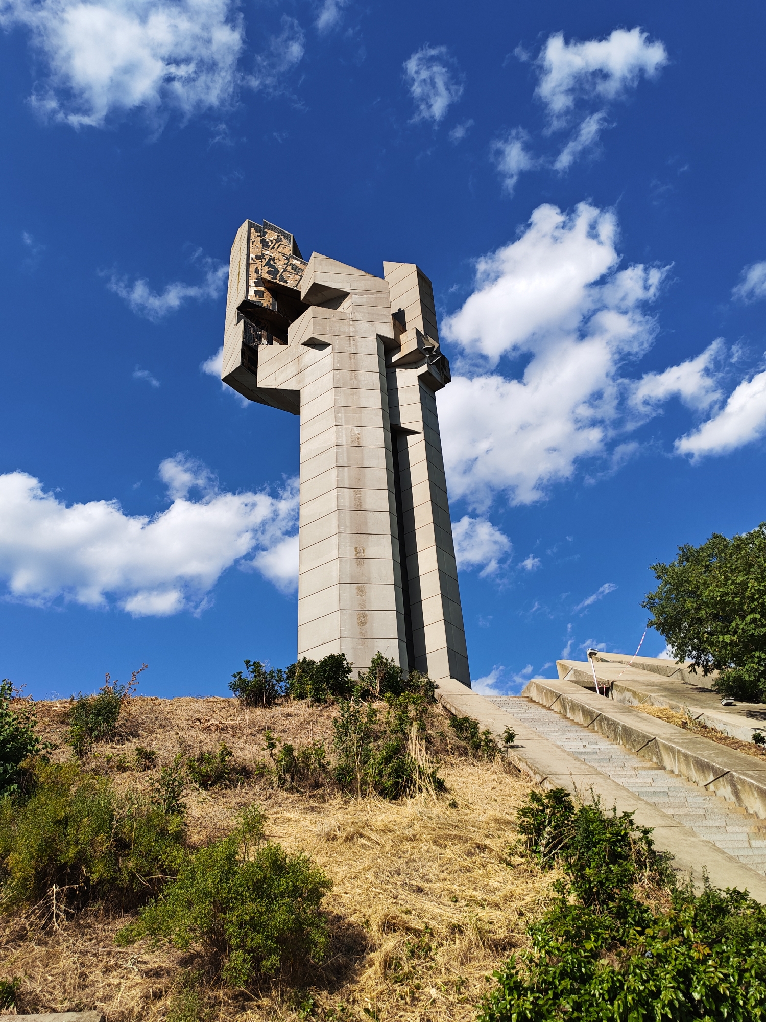 The image shows the Stara Zagora Defenders Monument, a concrete memorial structure located on a grassy hillside in Stara Zagora, Bulgaria. The monument is a tall, angular structure with multiple protruding sections and a cross-like shape at its upper portion. Concrete stairs ascend the hillside to the right of the monument, bordered by sparse vegetation and dry grass. The sky above is a vibrant blue, dotted with fluffy white clouds. The monument is the central subject, presented from a low-angle perspective. The environment appears to be an outdoor public space, likely a park or memorial site. There are no people visible in the image.