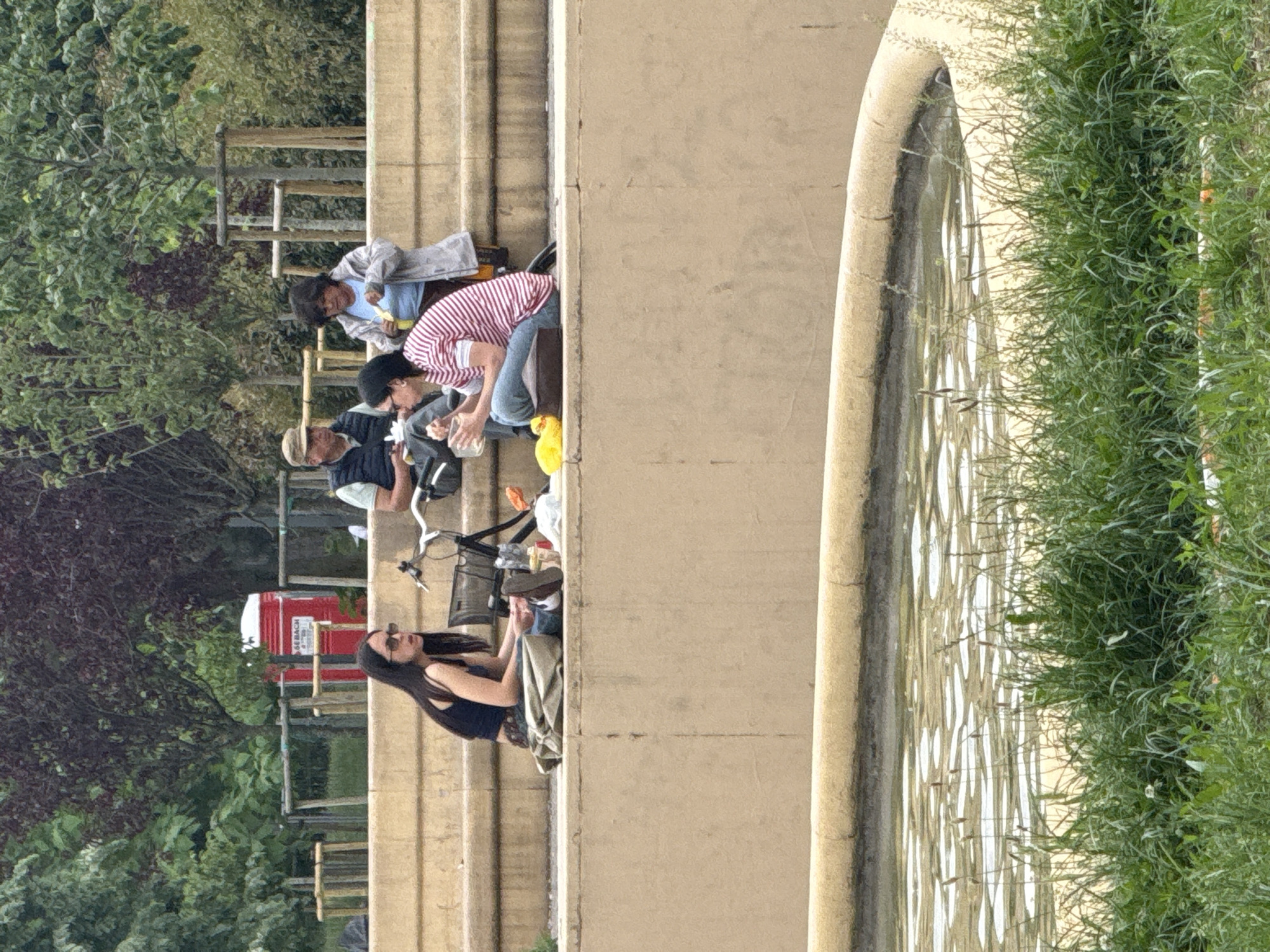 This outdoor scene, set in what is stated to be Milan, Italy, captures a relaxed gathering of several people on a sunny day. The image is oriented vertically, showing individuals seated on a broad, multi-tiered stone or concrete structure, possibly a wide set of steps or a low wall.
In the foreground, to the right, is a decorative circular water feature with a textured, possibly patterned, base visible beneath the water's surface. Lush green grass and vegetation grow densely along the very edge of this water feature.
The main subjects are a group of at least five people of various ages, casually dressed. They are sitting or stooping on the stone structure. One woman with long dark hair, wearing sunglasses and a dark top, is seated towards the bottom left, next to a bicycle. Further up, an older man with a light-colored hat is visible, alongside another person in a dark top and a person wearing a distinctive red and white striped shirt and a dark cap. This group appears to be sharing food or drinks, with plastic bottles and containers visible near them. Another individual in a light grey jacket and blue shirt is seated higher up the steps.
In the background, behind the people, are numerous green trees and bushes, indicating a park-like environment. A red portable toilet is visible amidst the greenery and a wooden fence, with partially legible text that includes "GEB. BACH." Two bicycles are present, suggesting some of the individuals arrived by bike. The overall atmosphere is one of leisure and outdoor enjoyment.